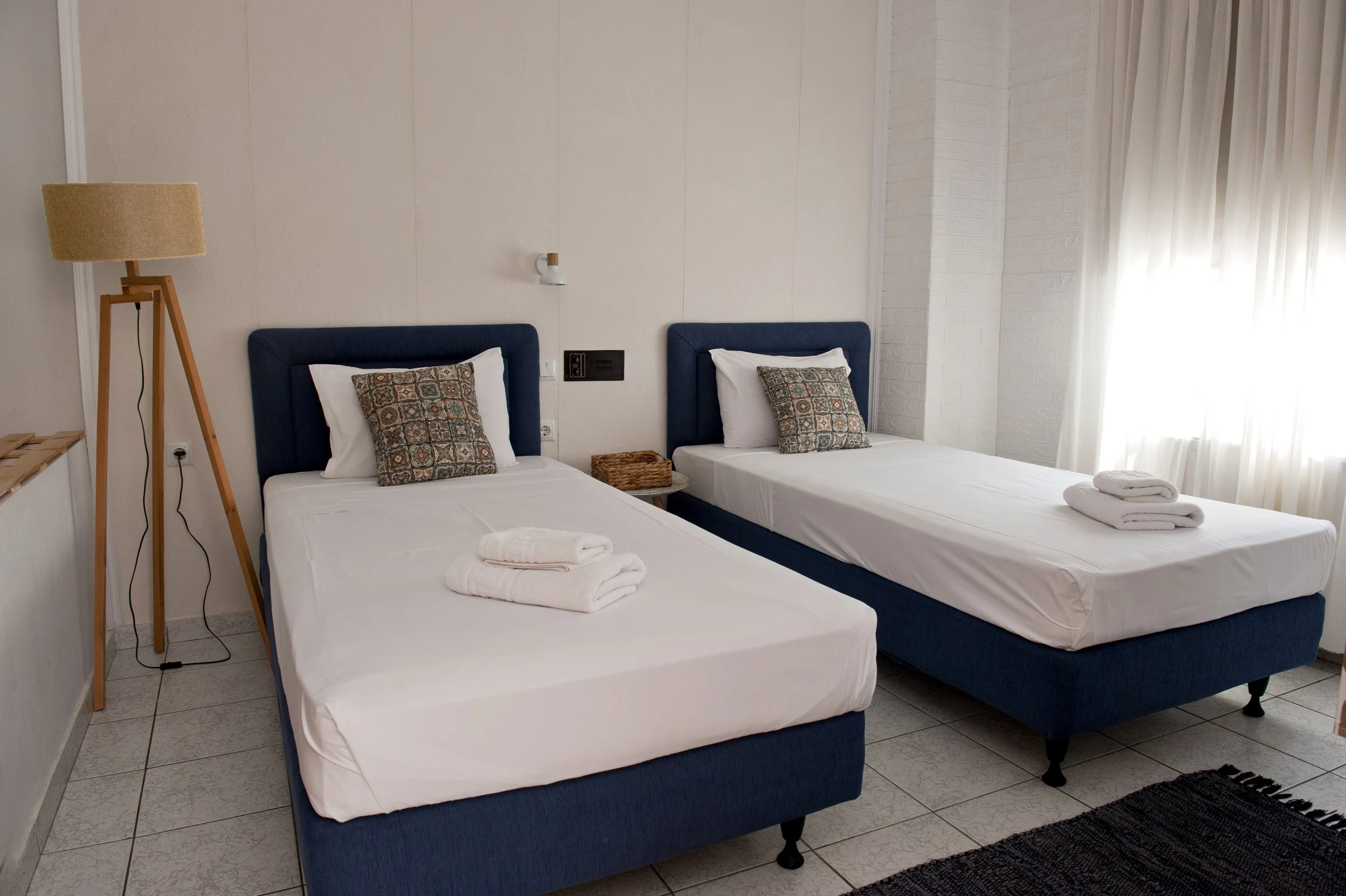 Two beds with white sheets and decorative pillows, located in a room with white walls, a wooden standing lamp, and a window with white curtains.
