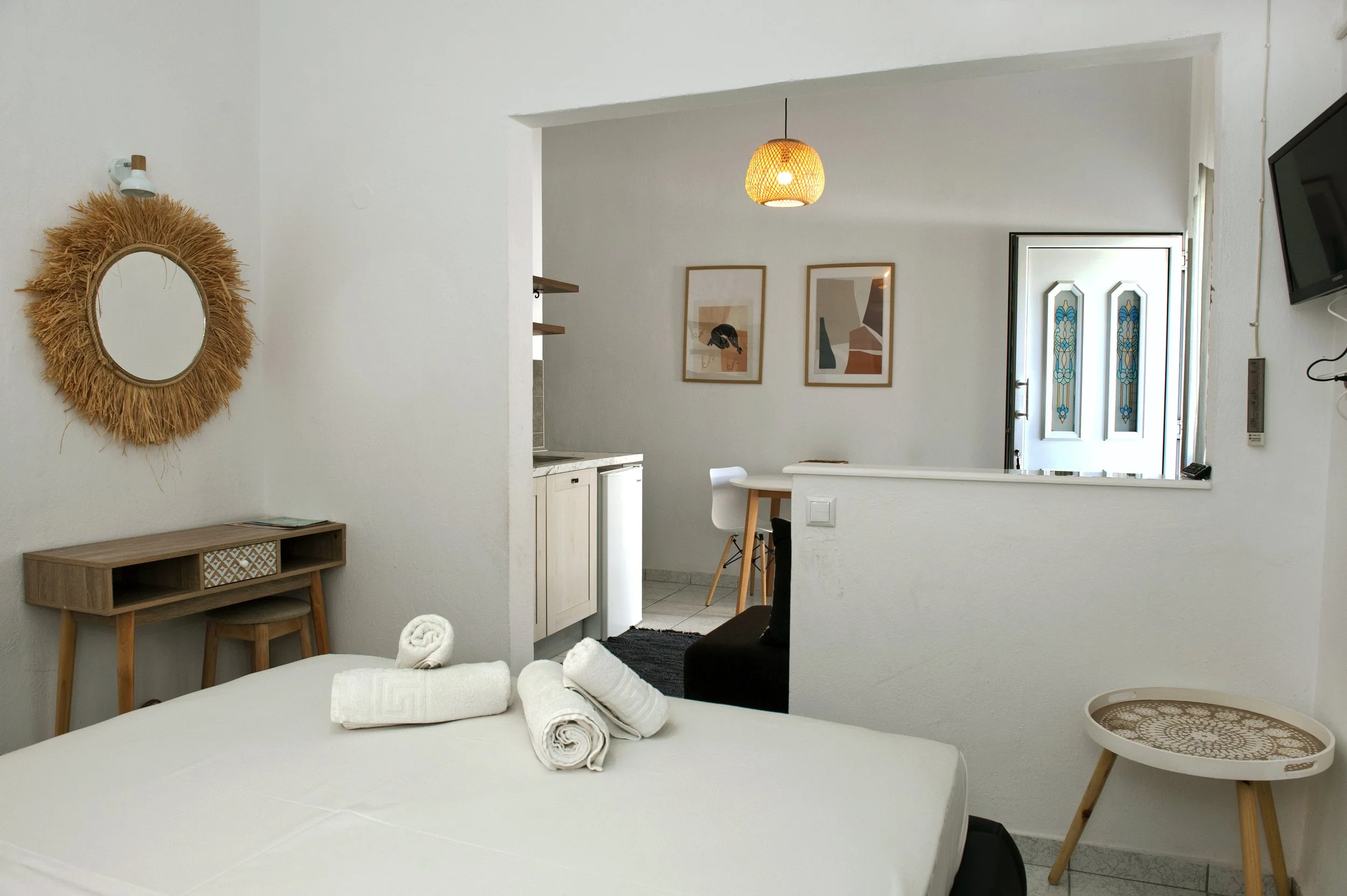 Hotel room with a bed, towels, and minimal decor, including a mirror with a straw frame, wall art, and a small table with matching stools.