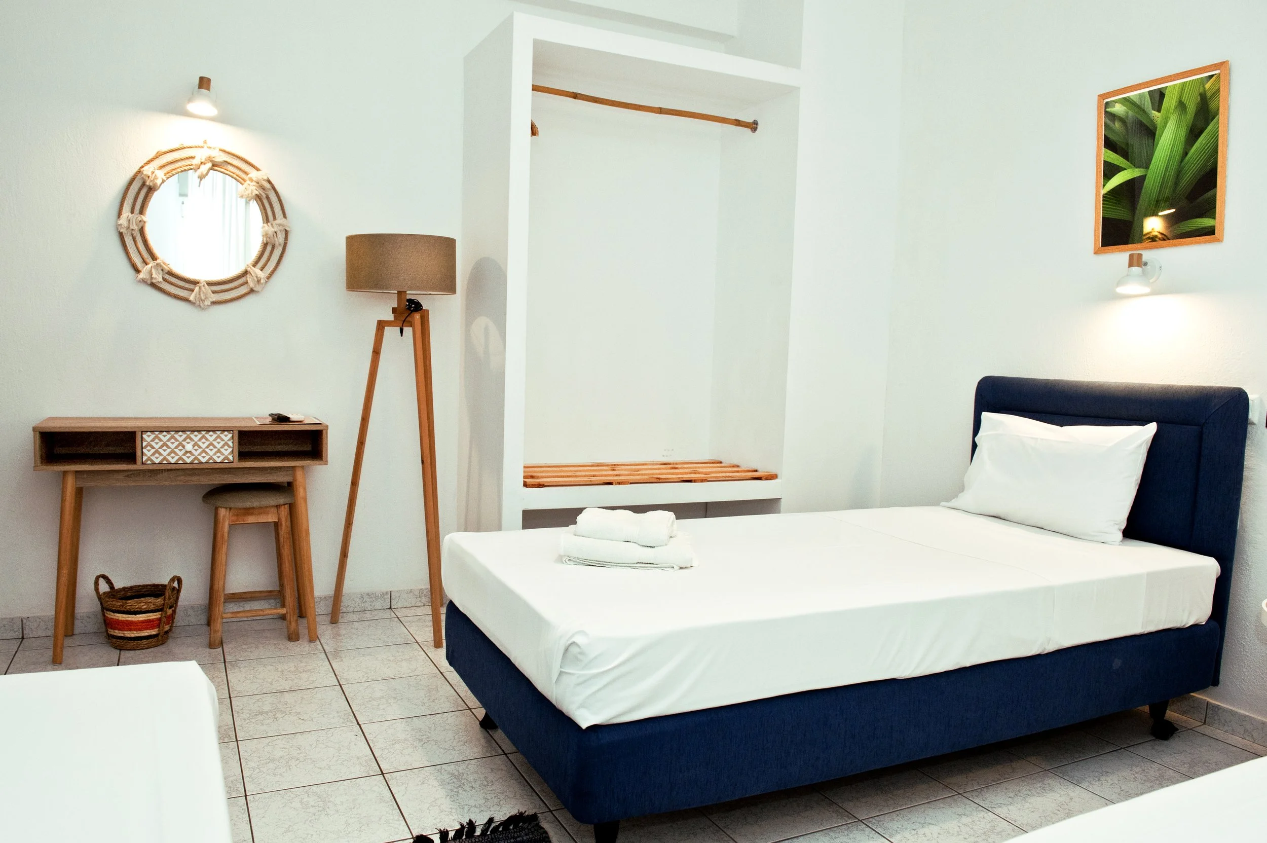 A hotel room with white walls, a bed with white linens and dark headboard, a wooden desk with patterned drawer and stool, a tall floor lamp, framed mirror with rope decor, and a mirror reflecting green plants.