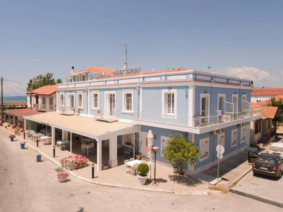 A light blue, three-story hotel with a sign reading "Hotel Kastero" on the roof. The hotel has a terrace on the second floor with white railings and outdoor furniture. There is an outdoor dining area with tables and chairs under a white canopy on the