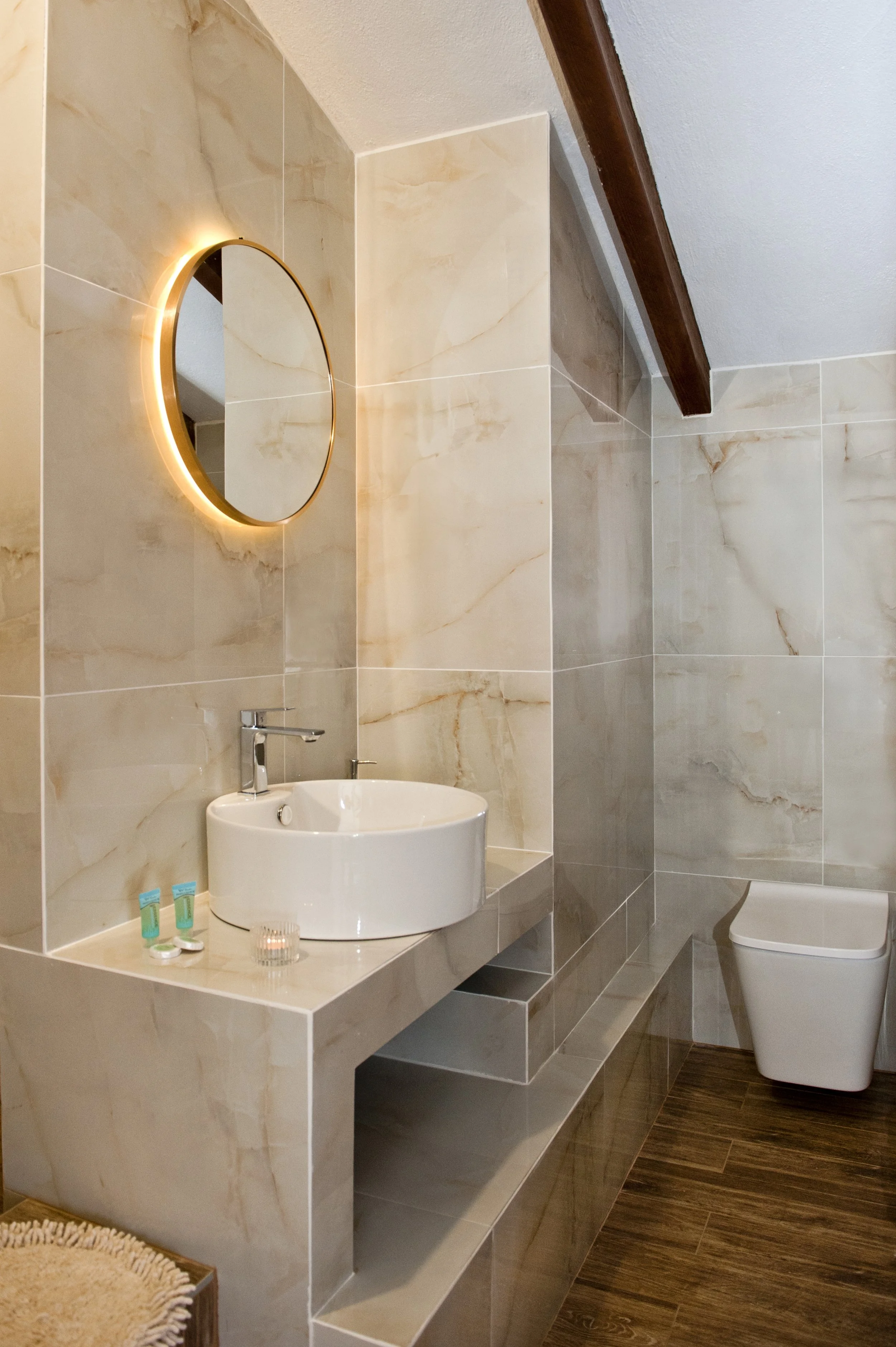 A modern bathroom with beige marble walls, a white round vessel sink, a circular mirror with backlit lighting, toiletries on the counter, and a toilet in the background. The space has wood accents and a clean, elegant design.