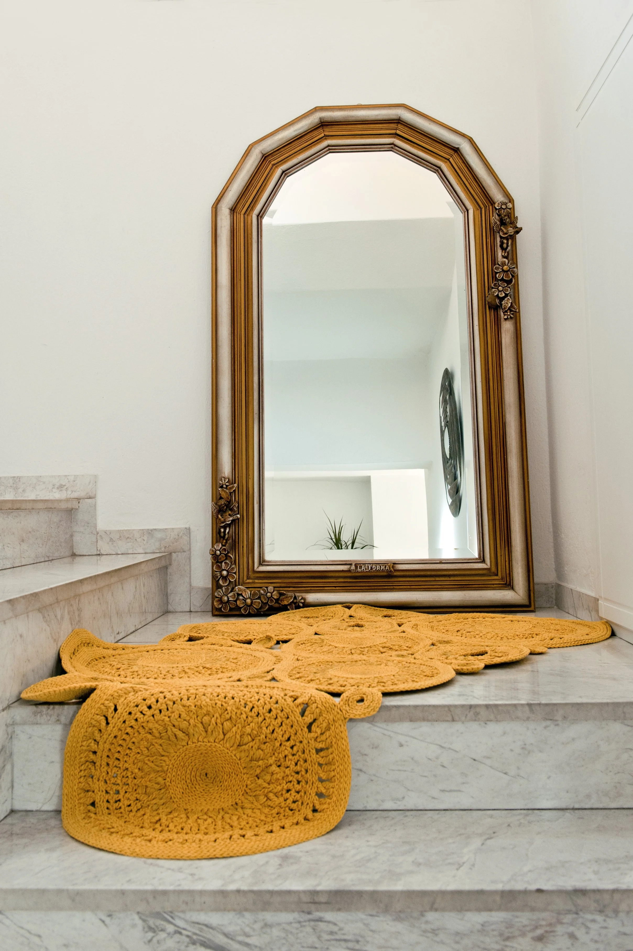 A large ornate gold-framed mirror leaning against a white wall above a marble staircase. A yellow crochet rug is on the stairs and a matching ottoman in front of the stairs.