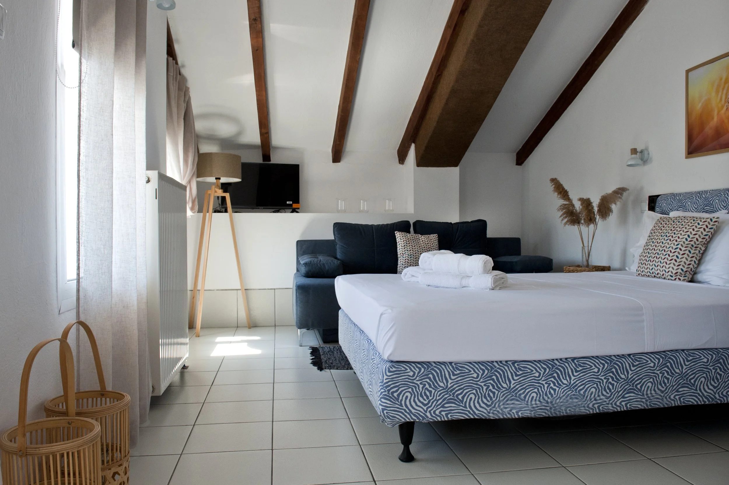 A bedroom with a bed covered in white linens, decorated with towels and pillows, a black sofa, a wooden lamp, a TV, a window with curtains, and a ceiling with exposed wooden beams.