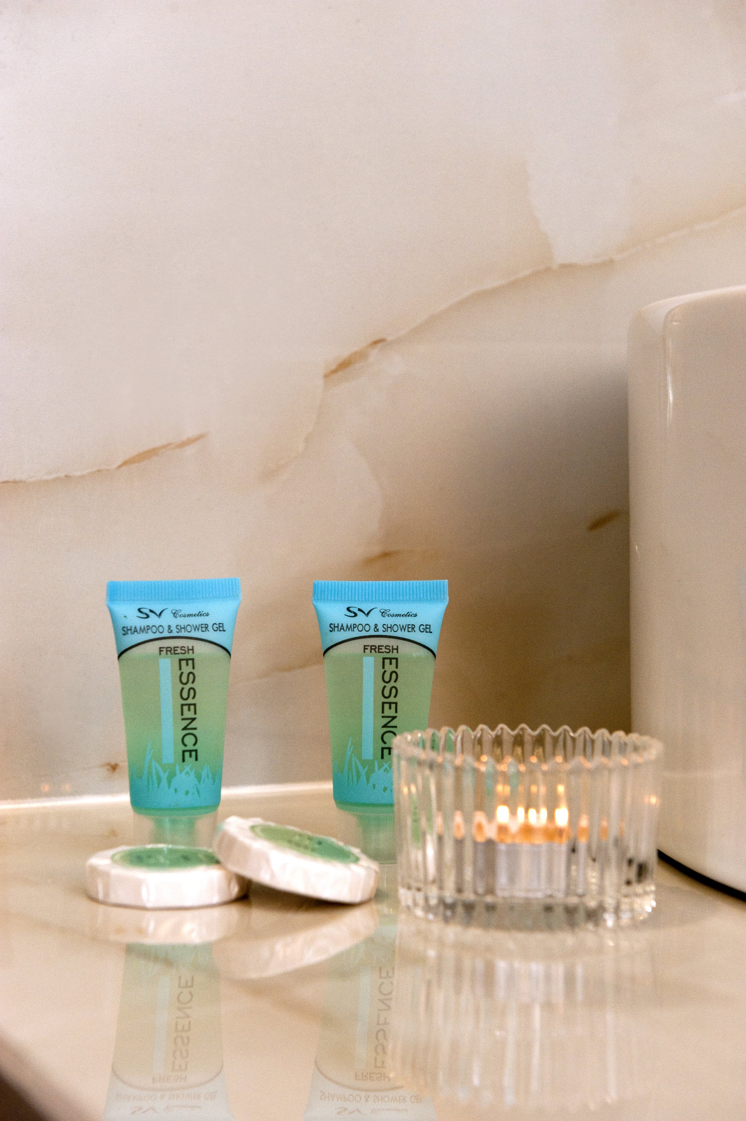 Two small tubes labeled 'SHAMPOO & SHOWER GEL' in blue and green, a glass candle holder with a lit candle, and a white container with green contents on a reflective surface in front of a cracked beige wall.