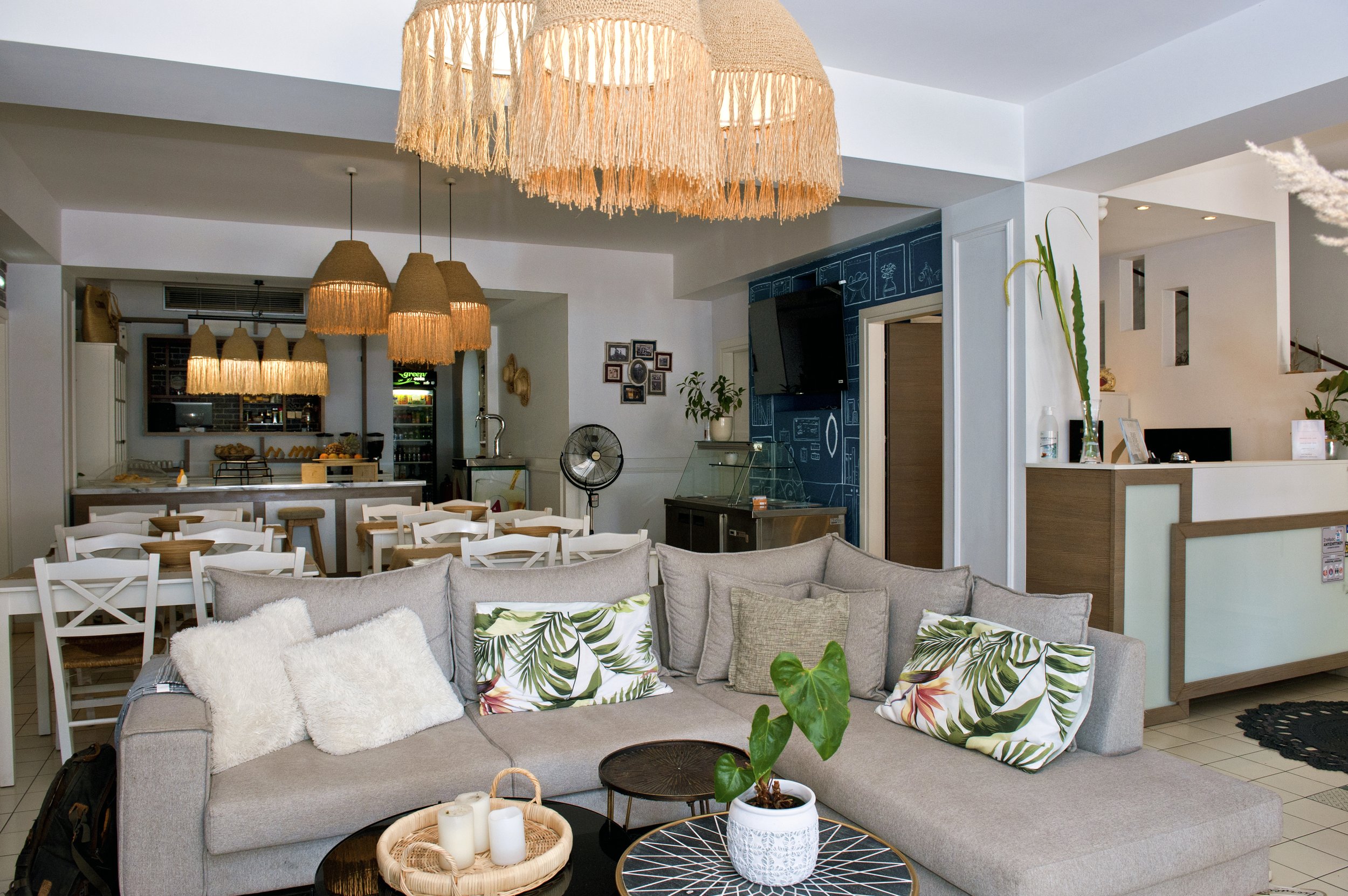 A cozy living room with a gray sofa decorated with patterned and plain pillows, a black wireframe coffee table with a white pot and candles, and a potted plant in the foreground. Behind the sofa, there is a dining area with white chairs, a kitchen wi