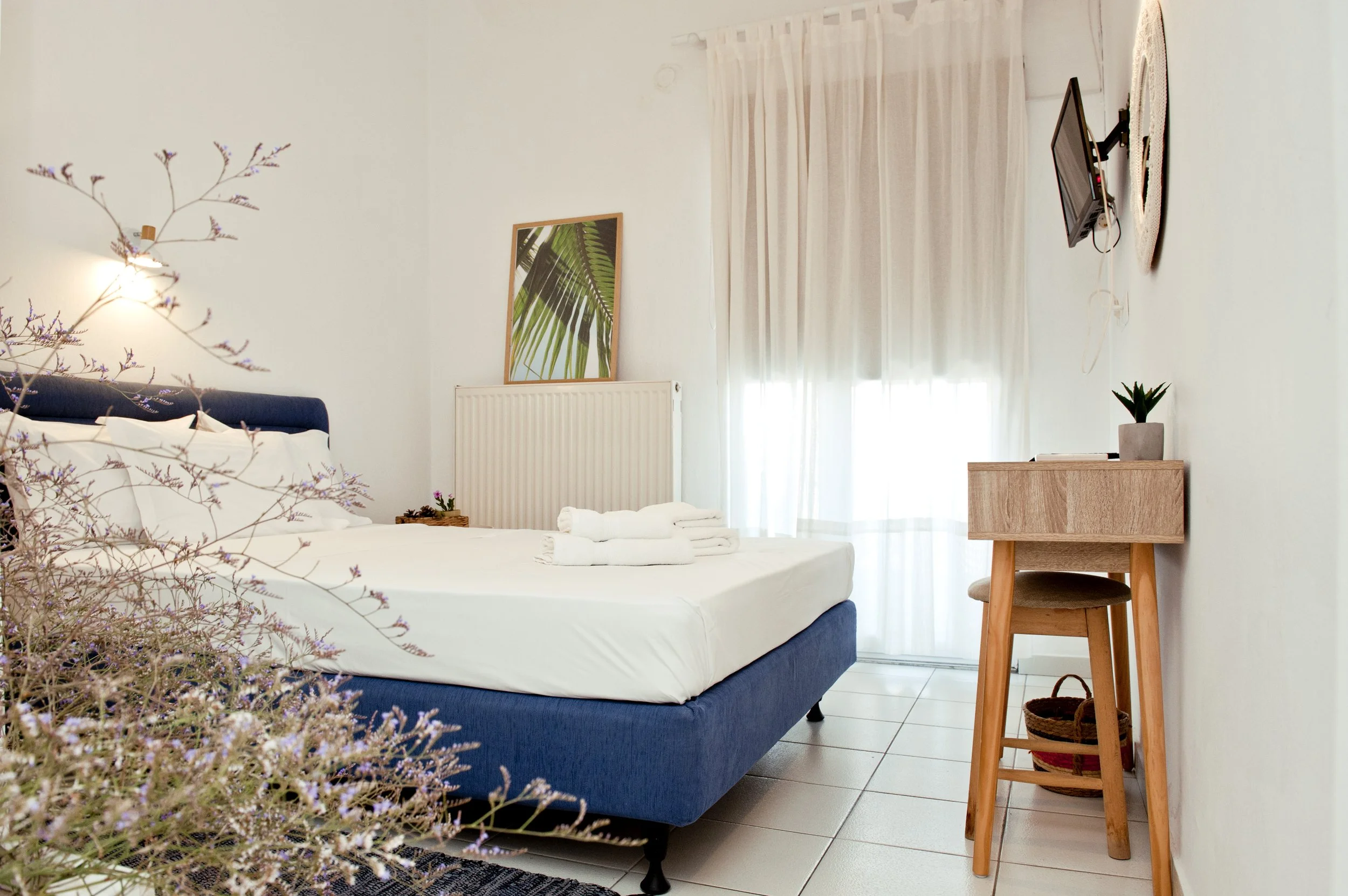 A bedroom with a white bed and blue headboard, a window with light curtains, a framed green leaf print on the wall, a small wooden desk with a potted plant, a TV mounted on the wall, and a flower arrangement in the foreground.