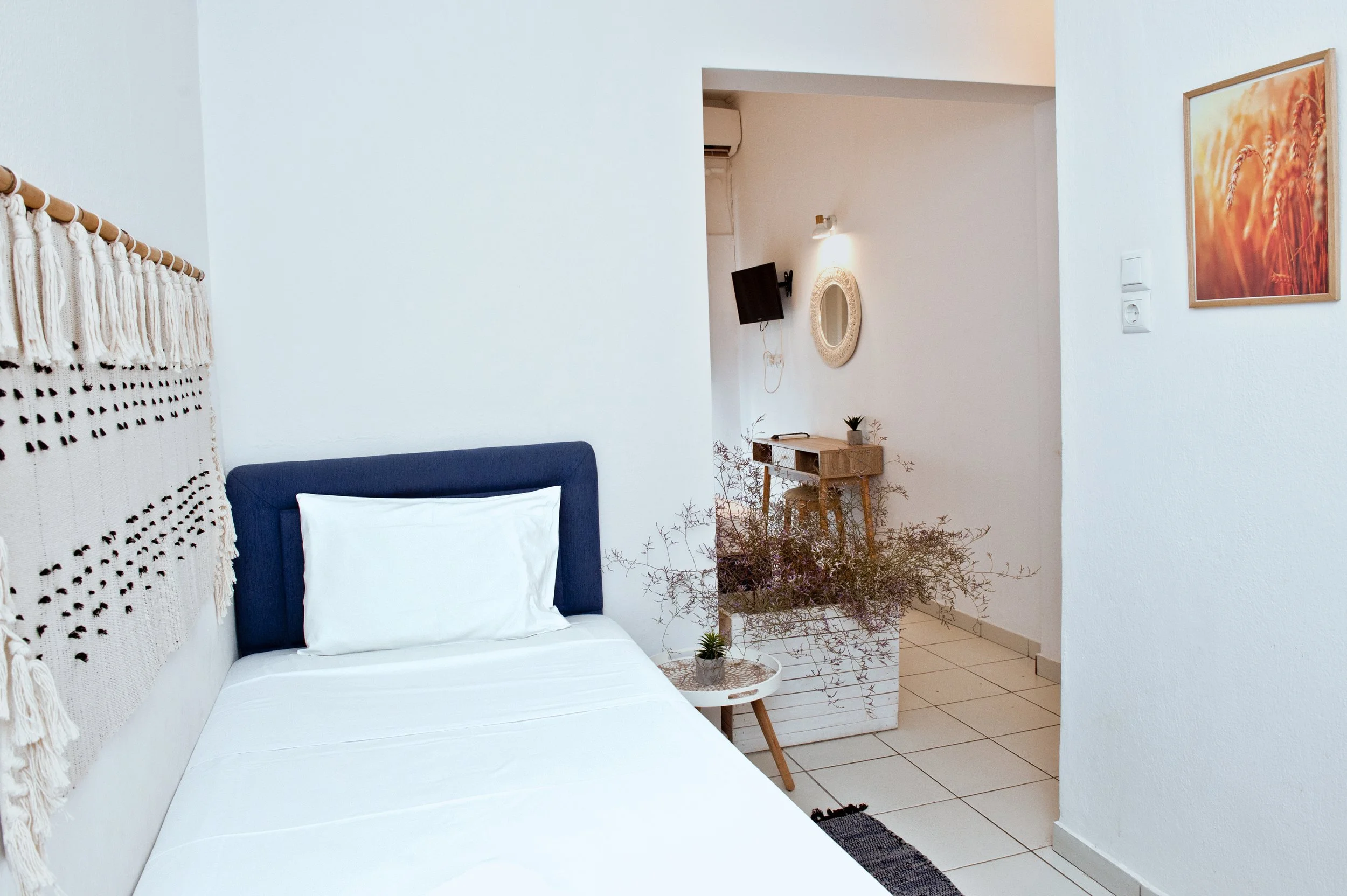 A cozy bedroom with a bed featuring a navy headboard and white linens, a black wall-mounted TV, a small wooden side table with potted plants, wall art, and a decorative wall hanging.