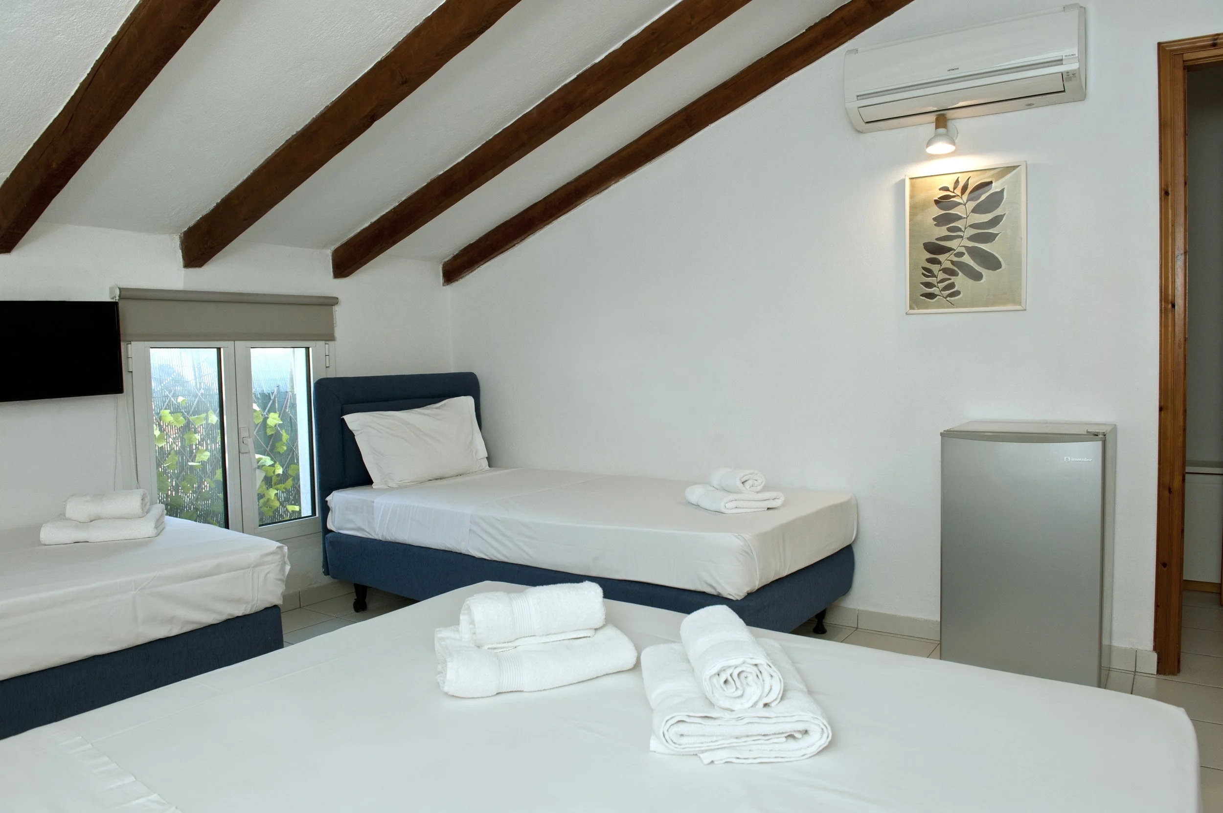 A cozy bedroom with sloped ceiling and exposed wooden beams, featuring three beds with white linens and rolled towels, a small refrigerator, a wall-mounted TV, a window with a view of greenery, and wall art of leaves, in a minimalist style.