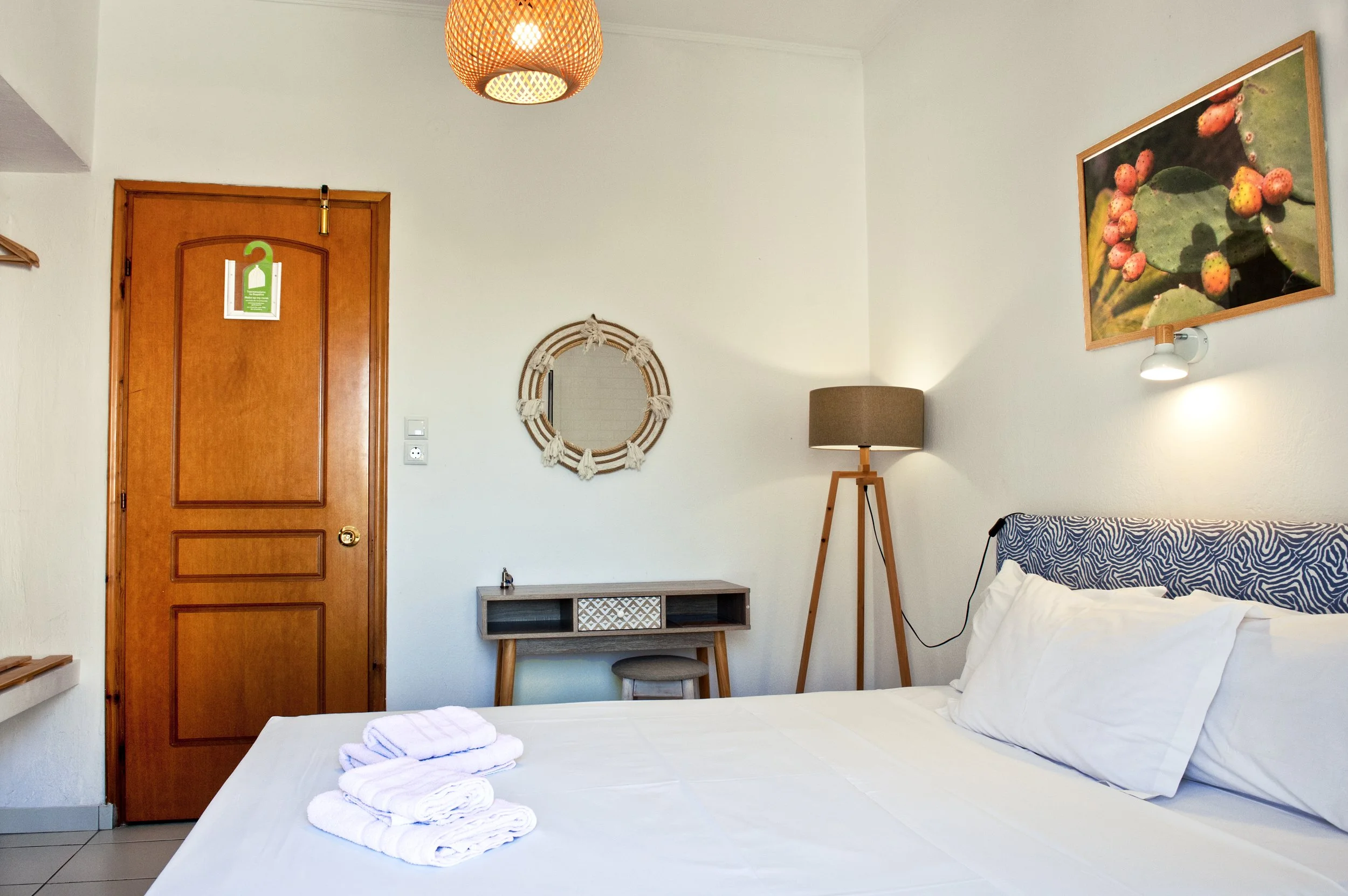 A bedroom with a wooden door, wall art of strawberries, a standing lamp, a small table, and a bed with rolled towels on it.