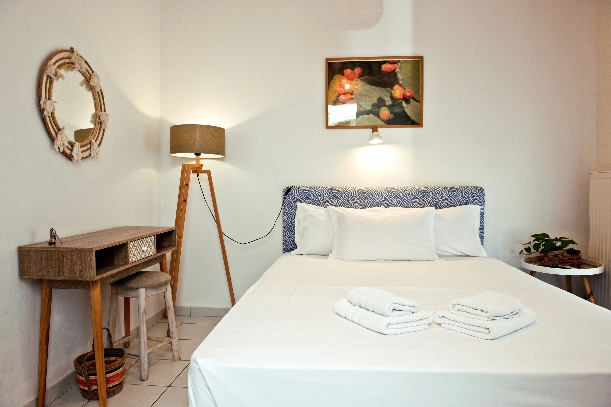 A neatly made bed with a white sheet and pillows, two towels folded on top, a small wooden nightstand with a plant, a modern tripod floor lamp, a framed botanical picture with red berries, a decorative oval mirror with tassels, and a stool, all in a 