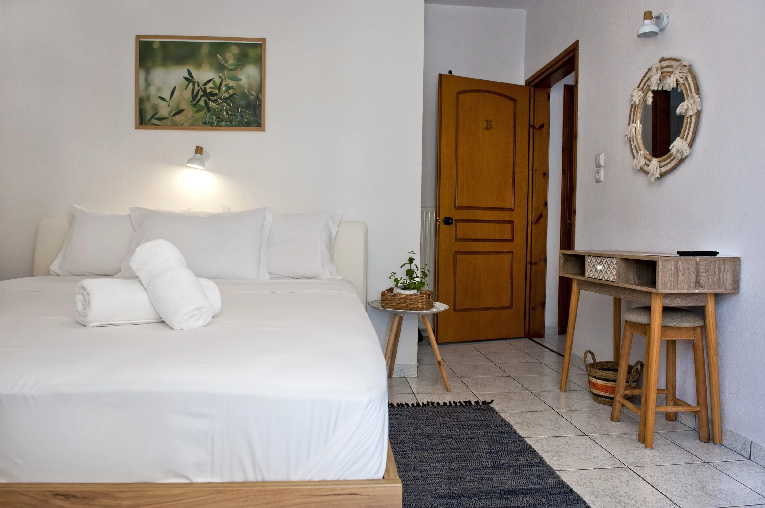 A bedroom with a white bed, two rolled towels, a small wooden side table with a plant, a wooden door with the number 15, a framed picture of greenery on the wall, a wall light, a decorative mirror with a ruffled border, a small wooden desk with a cha