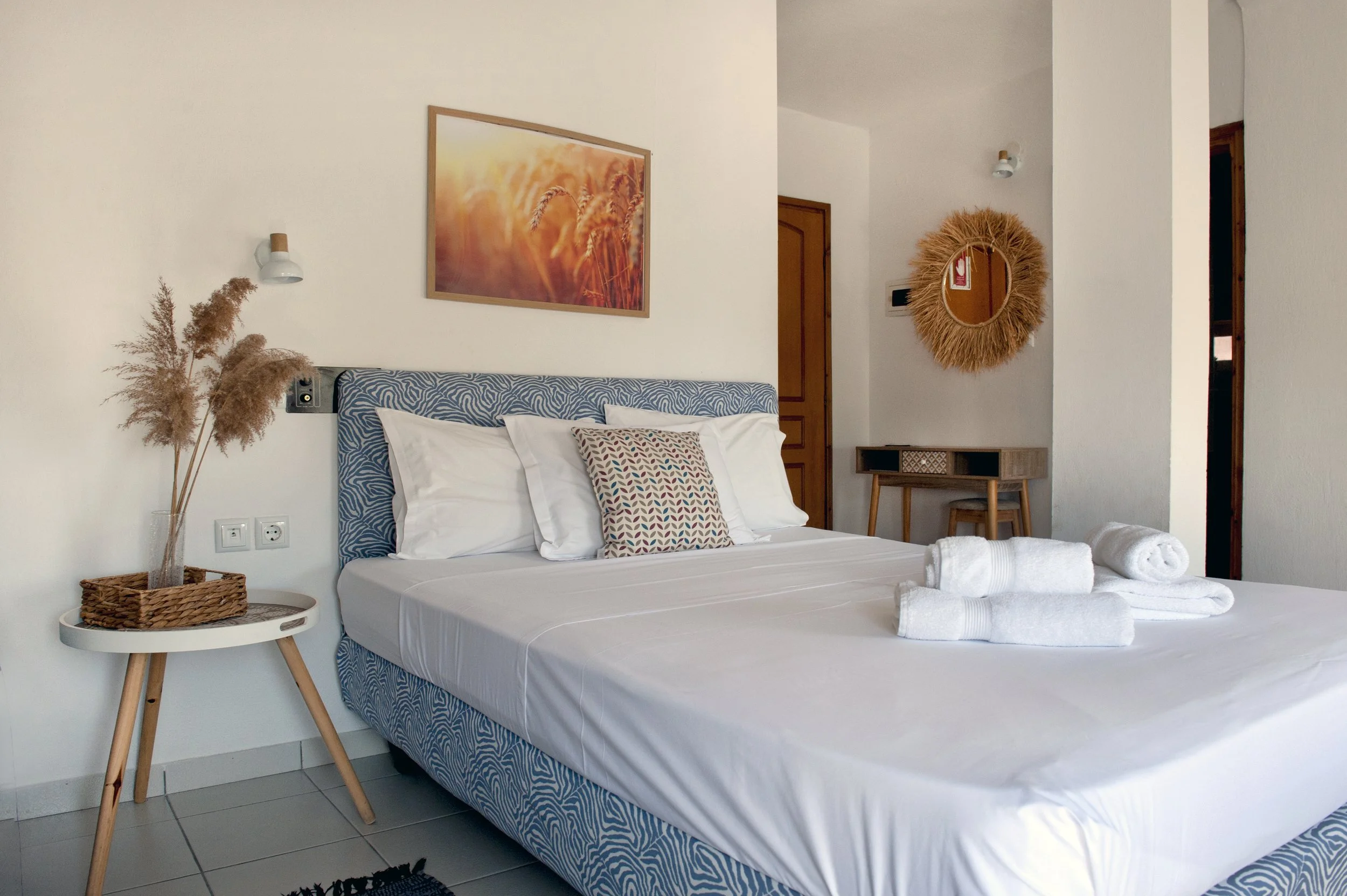 Hotel room with a bed featuring a patterned blue headboard, white linen, and pillows, a side table with dried plants in a glass vase, wall-mounted reading lights, a framed sunset-themed artwork, a round mirror with a straw frame, and a small wooden d