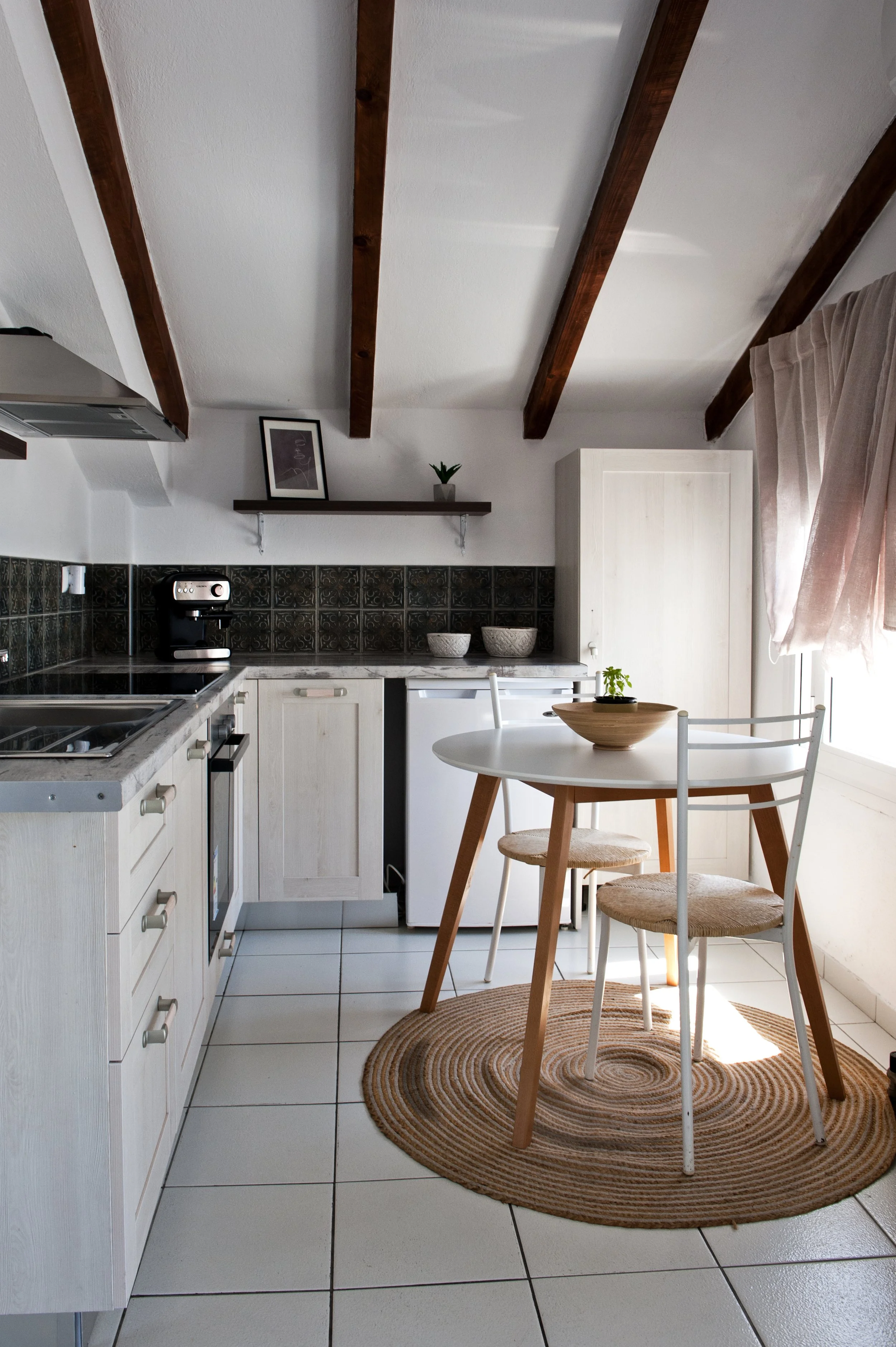 A cozy kitchen with white cabinetry, wooden beams on the ceiling, a round dining table with three chairs, a small potted plant, framed artwork, a coffee machine, bowls, and a woven circular rug.