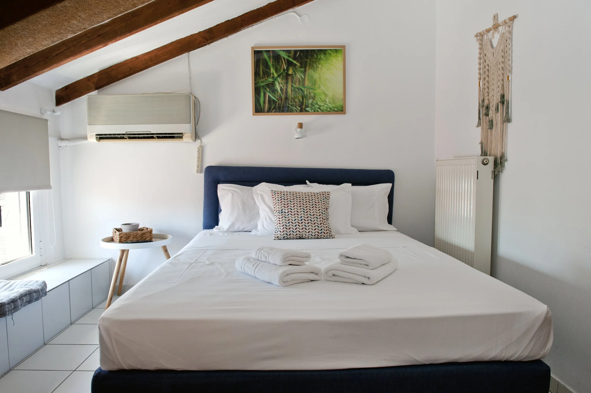 A neatly made bed with white linens and four folded towels on top. The bed has a navy headboard and a decorative pillow with a geometric pattern. To the left, there is a small round table with a basket and some items inside. Behind the bed, there is 