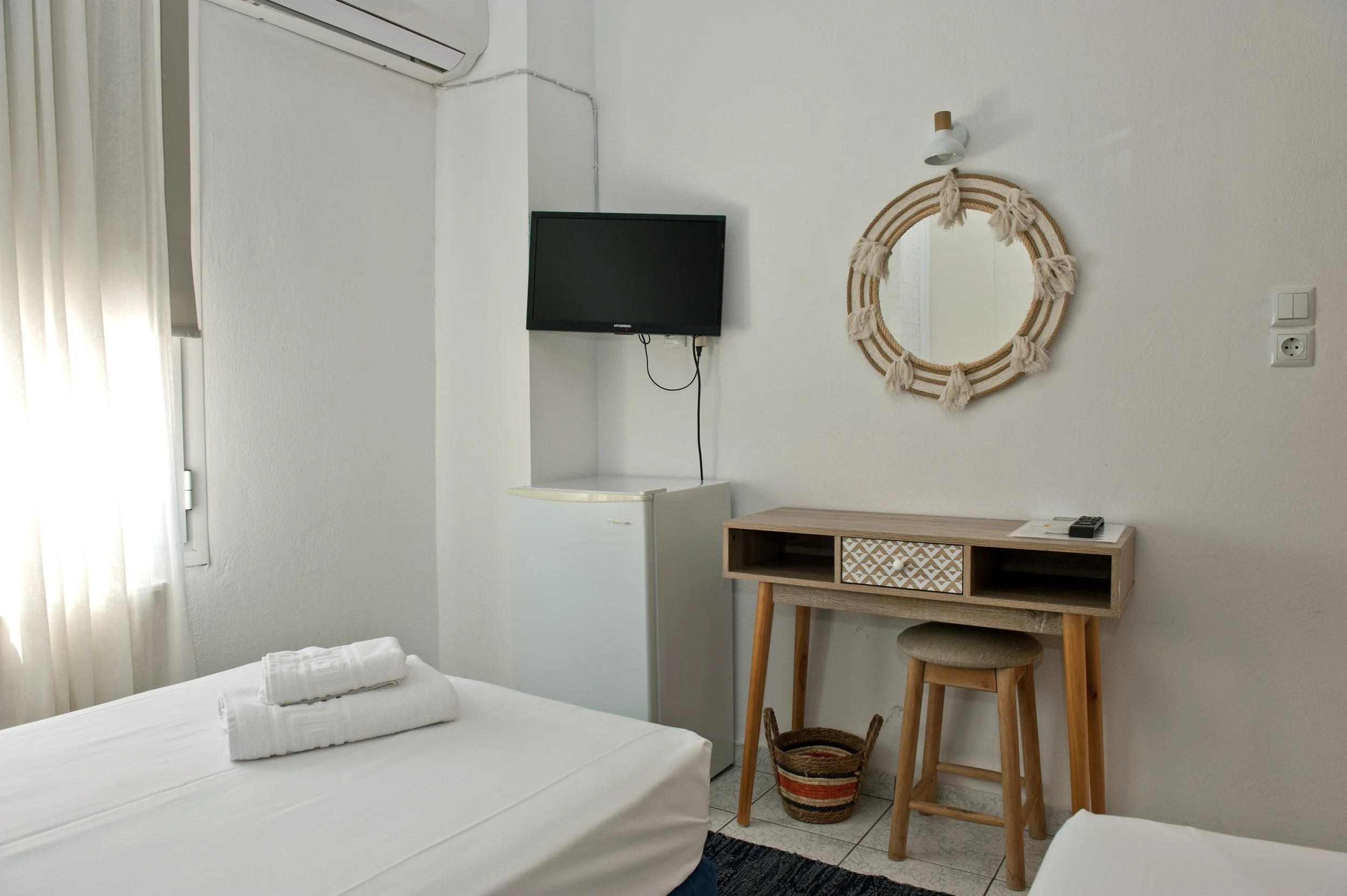 A minimalist hotel room with white walls and bed sheets, a small wall-mounted TV, a mirror with a woven frame, a wooden desk with drawers, a stool, and towels on the bed.