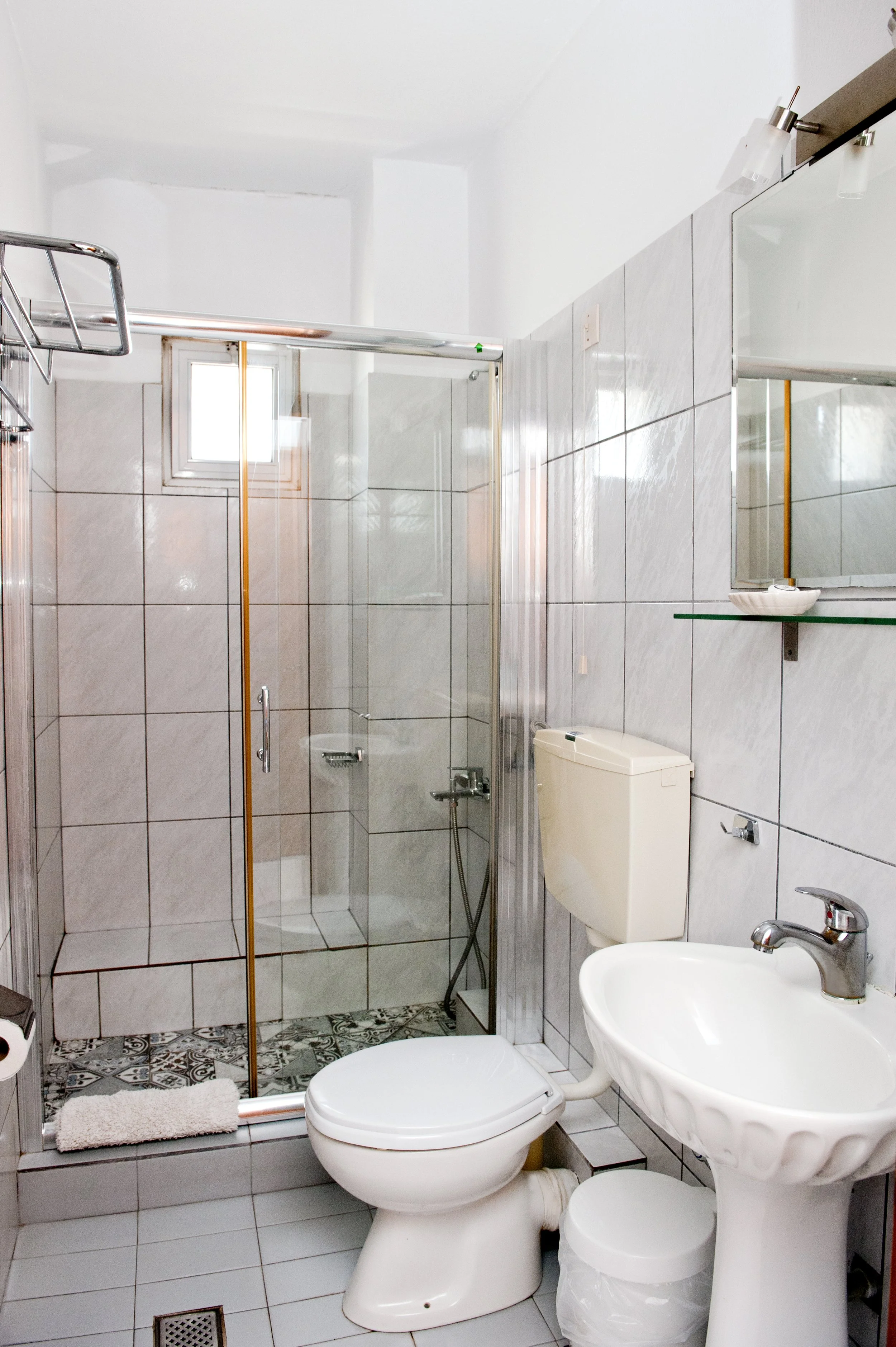 A small bathroom with a shower enclosure, toilet, and pedestal sink, tiled walls and patterned floor tiles, and a small window near the ceiling.
