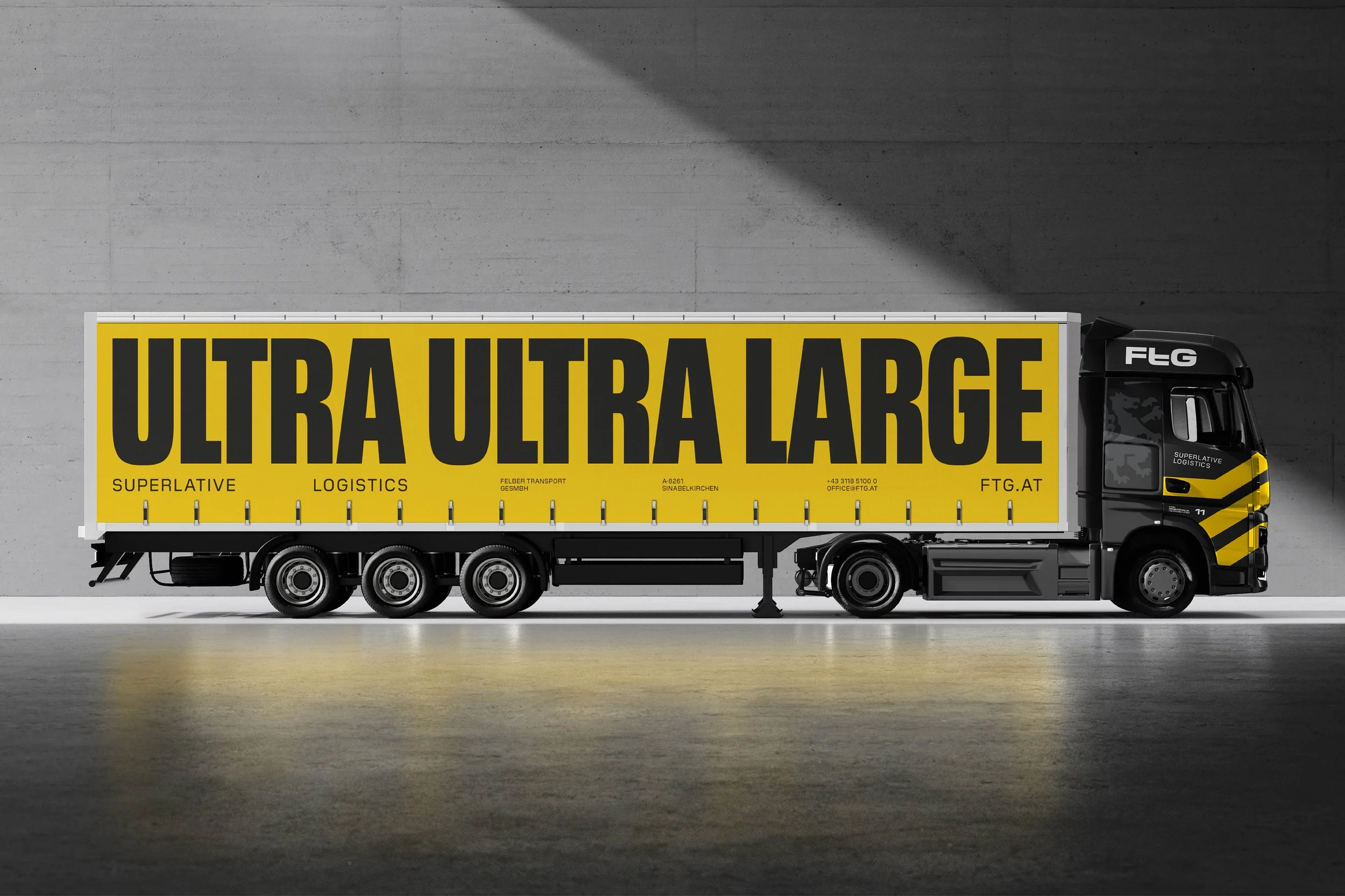 Großer LKW mit gelbem Schriftzug 'ULTRA ULTRA LARGE' auf der Seite, steht vor einer grauen Wand, trägt ein schwarzes Fahrerhaus mit Logo 'FTG' und 'SUPERLATIVE LOGISTICS'