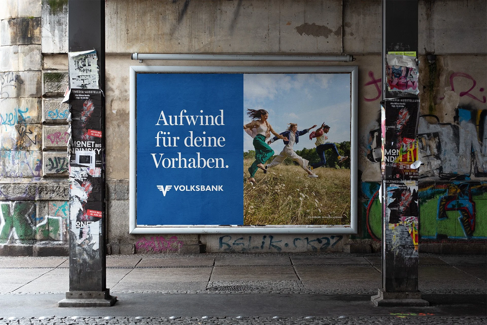 Werbung für Volksbank mit einem Bild von drei Menschen, die im Freien laufen, vor einem blauen Himmel und grünen Bäumen, auf einer gut frequentierten Straßenunterführung mit grauer, Graffiti-beschmierter Wand.
