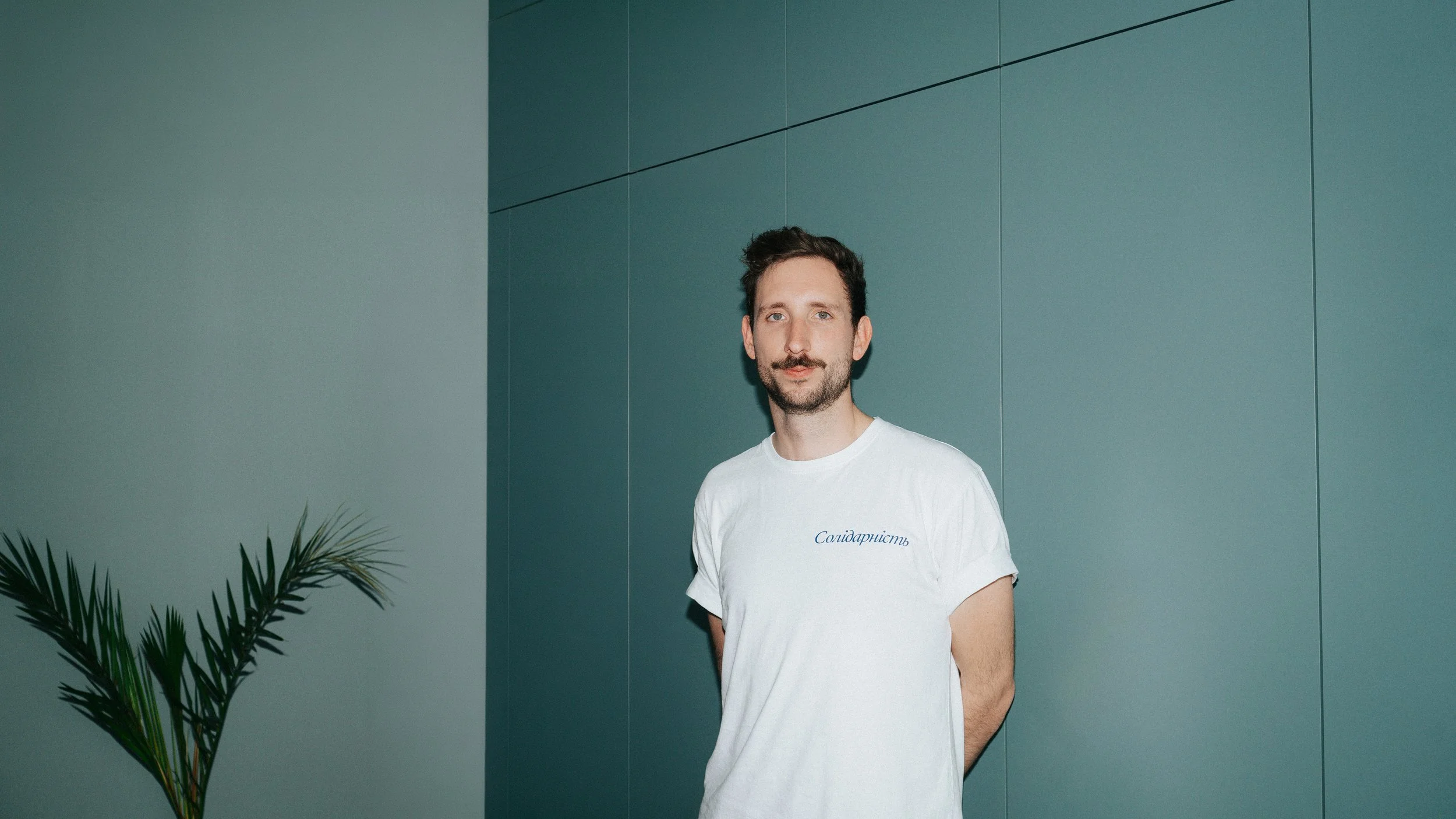 Ein Mann mit dunklem Haar und Bart, trägt ein weißes T-Shirt mit französischem Schriftzug, steht vor einer blauen Wand, neben einer Palme.