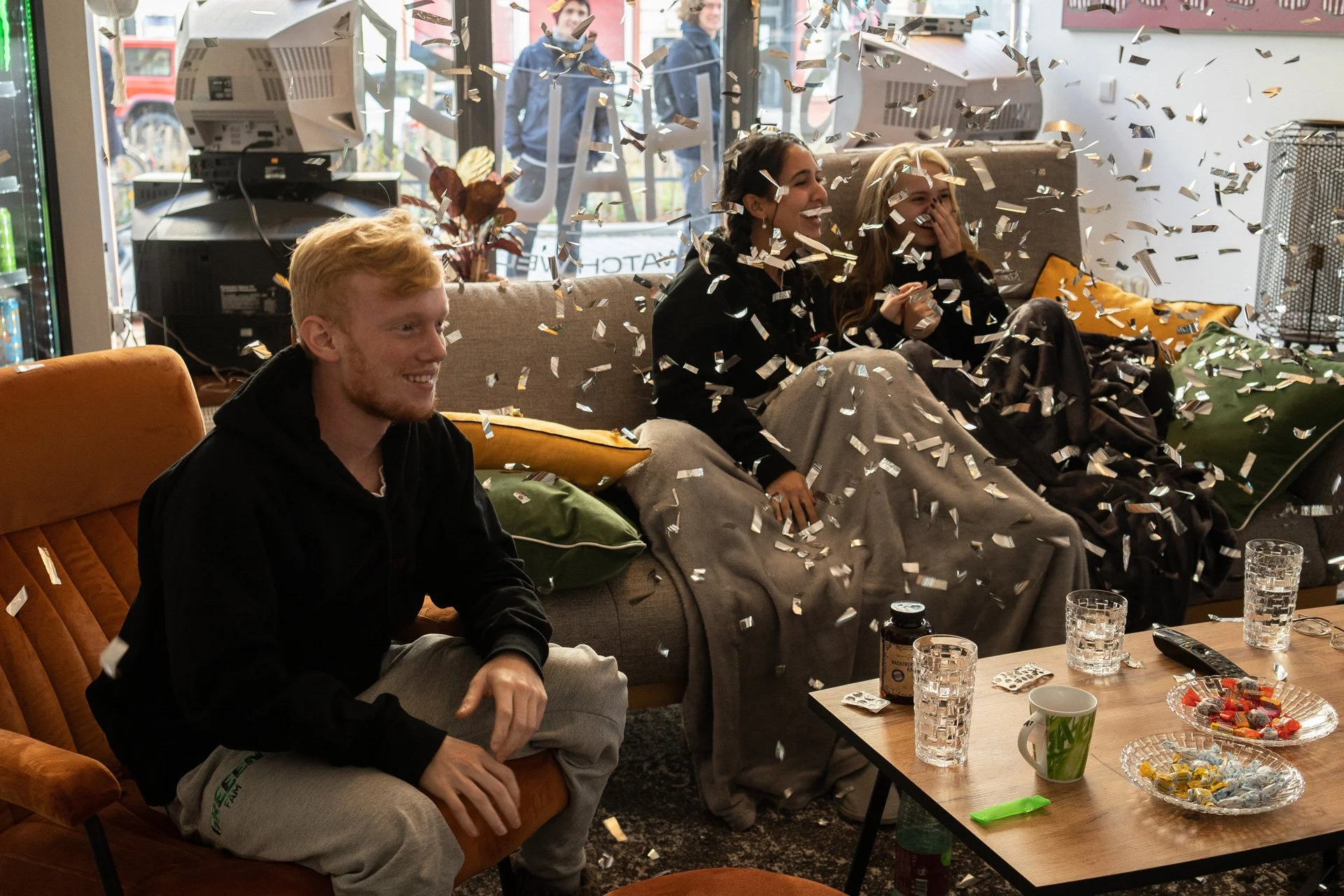 Gruppe von drei jungen Erwachsenen, die auf einem Sofa im Wohnzimmer sitzen und eine Feier mit Konfetti haben.
