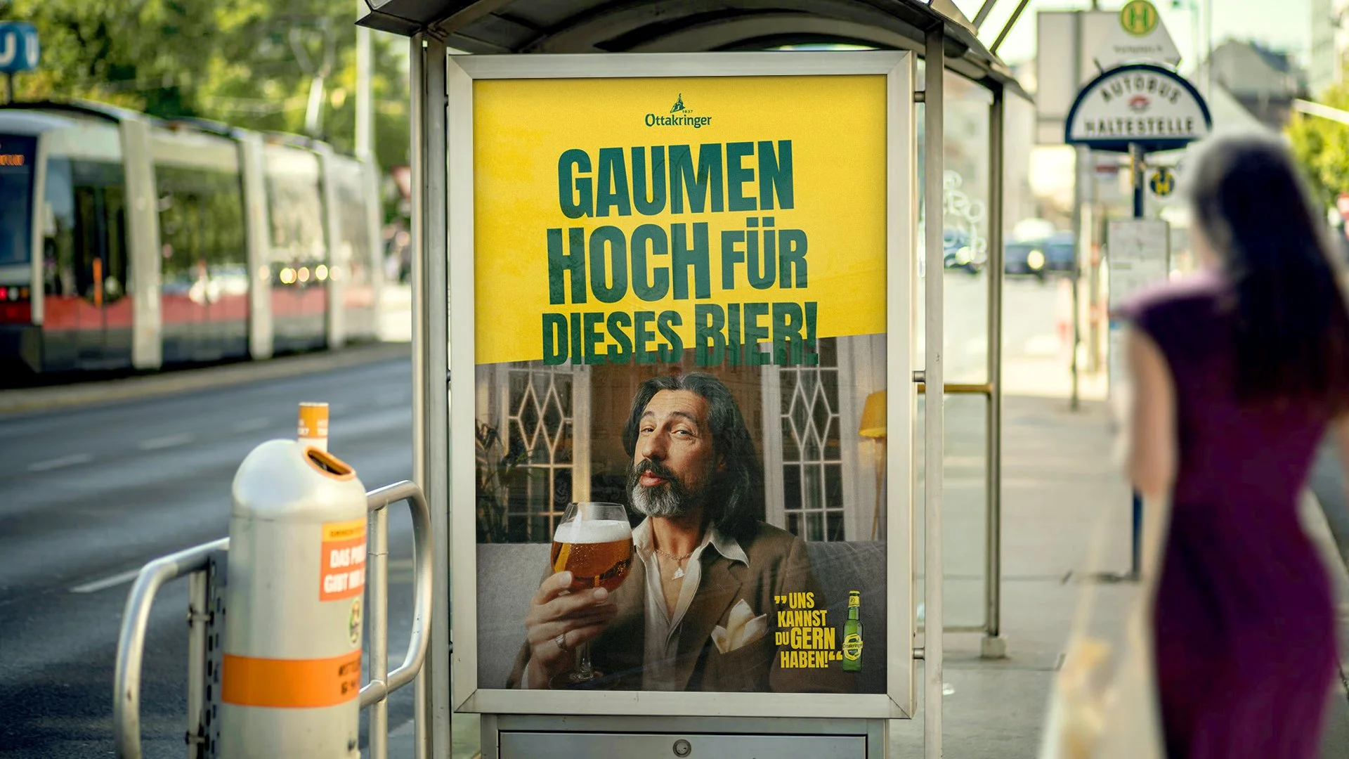 Ein Bushaltestellen-Werbeplakat mit einem Mann, der ein Bierglas in der Hand hält, und Text, der auf Hochprozentiges für dieses Bier hinweist. Im Hintergrund sind eine Straßenbahn, eine Frau und Verkehrsschilder zu sehen.