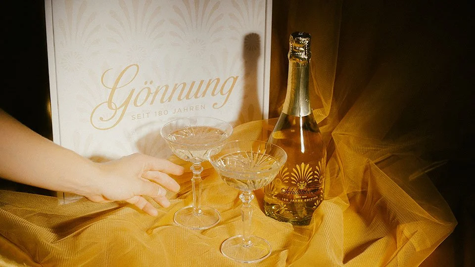 Zwei stilvolle Cocktailgläser auf einer goldfarbenen Stoffdecke neben einer Flasche Sekt mit Goldfolie und einer weißen Karte mit Goldschrift. Das Ganze ist festlich dekoriert.