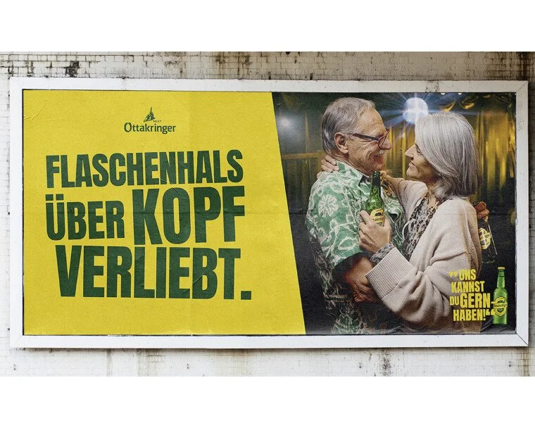 Werbung für Ottakringer Bier mit einem älteren Mann und einer älteren Frau, die tanzen und Bier trinken, auf einem Plakat an einer Wand. Die Headline sagt: "Flaschenhals über Kopf verliebt."