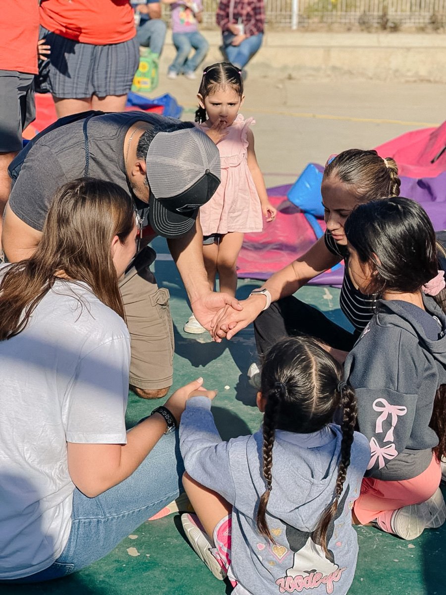 A group of people, including children and an adult, are gathered in a circle outside on a sunny day, holding hands and praying or having a moment of connection.