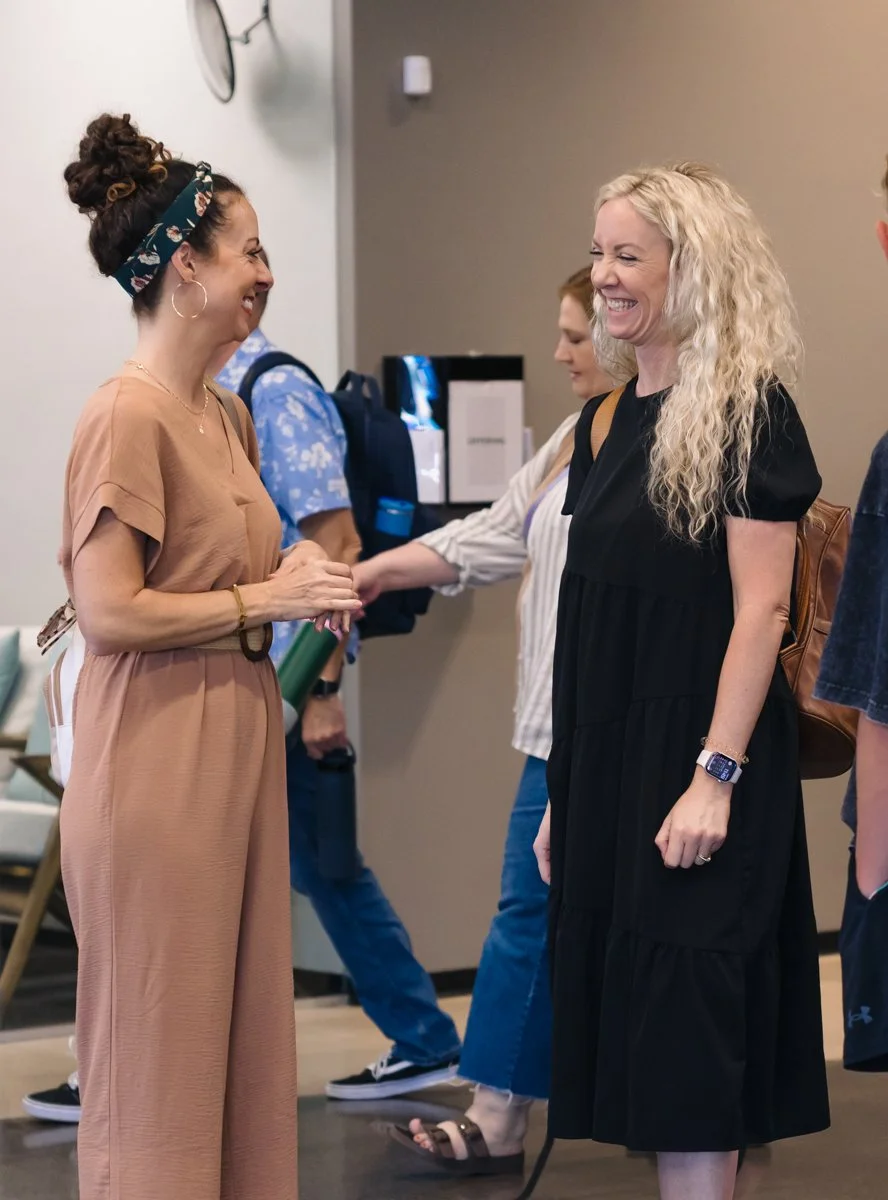 Two women smiling and talking to each other in a room with other people in the background.