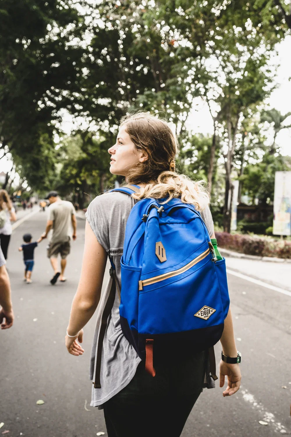 Young woman with a blue backpack walking in a park filled with green trees, with other people and children in the background.