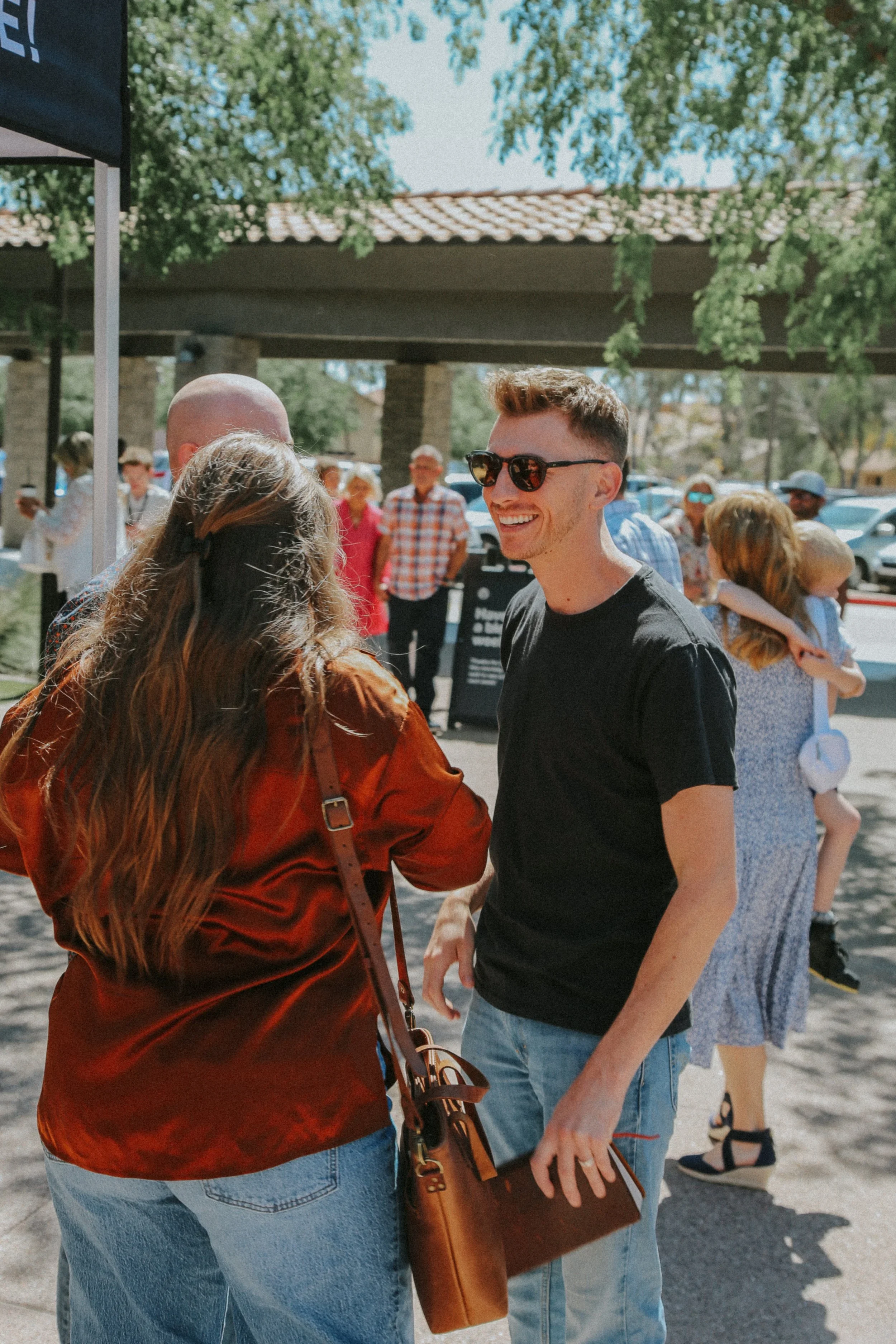 Two people, a young woman with long wavy hair and a young man with short hair and sunglasses, are smiling and talking to each other outdoors on a sunny day. Other people are seen in the background near parked cars and under a tree.