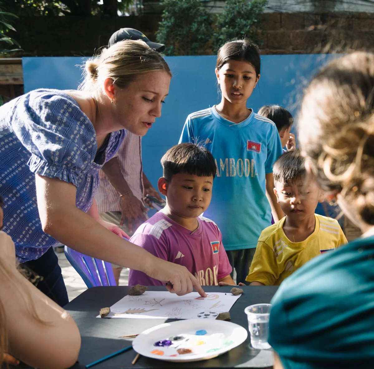 A woman and several children gather around a table outdoors, looking at a drawing on paper, with art supplies and rocks on the table, engaging in a group activity.