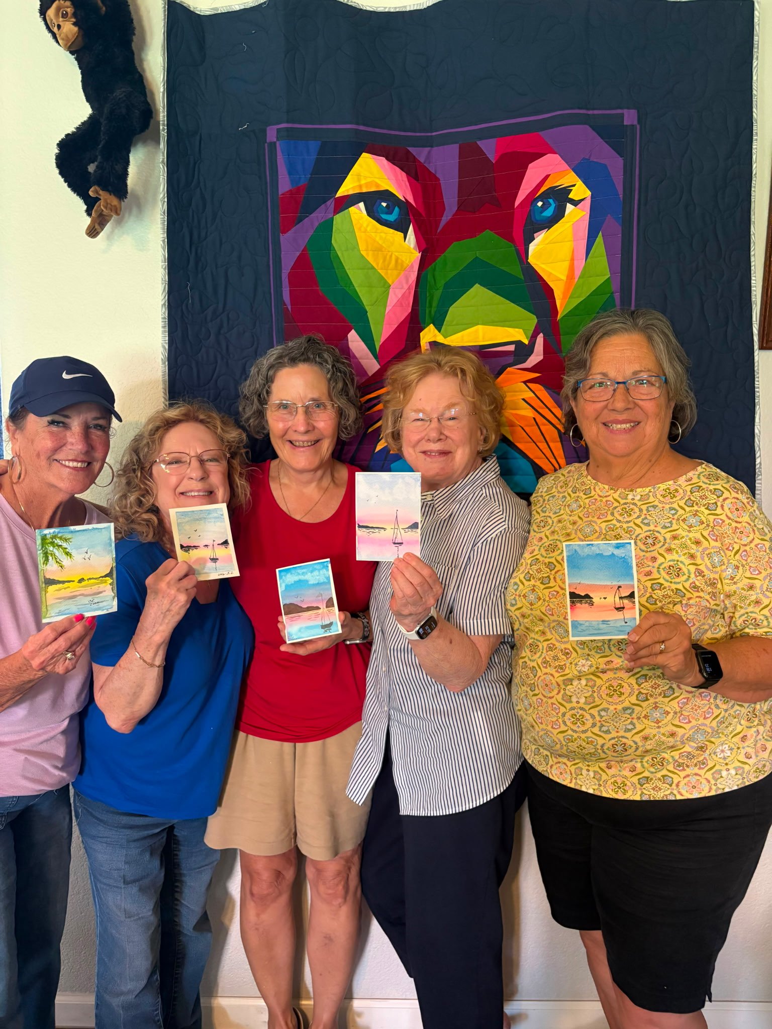 Group of four women standing in front of a colorful quilt with a dog face design on it. They are holding small paintings of landscapes and smiling.