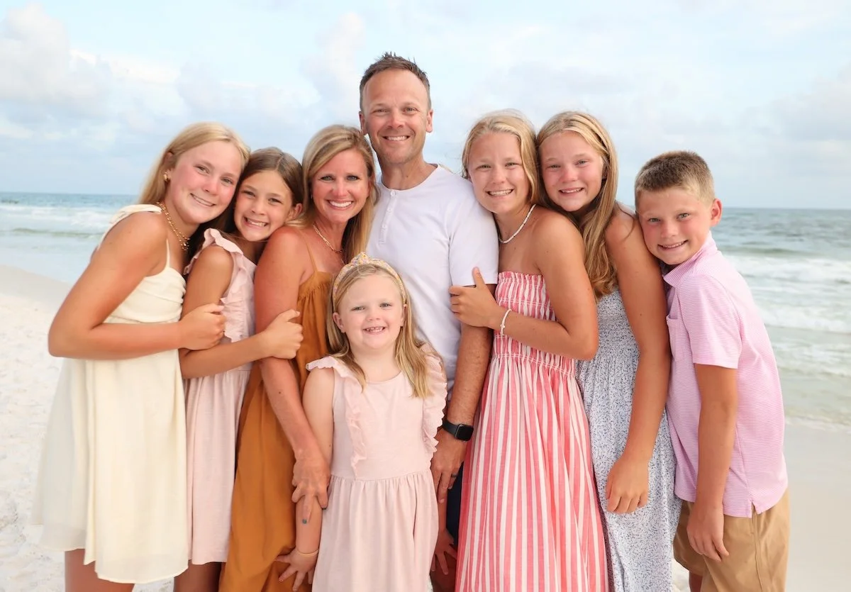 A family of nine standing on a beach, smiling at the camera with the ocean and sky in the background.