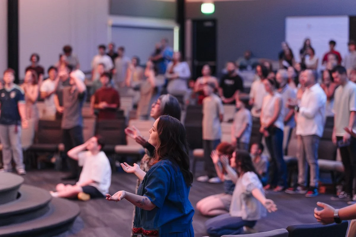 A group of people gathered in a dimly lit indoor space, some standing while others sit or kneel, appearing to participate in a collective prayer or meditation with their eyes closed and hands raised or folded.