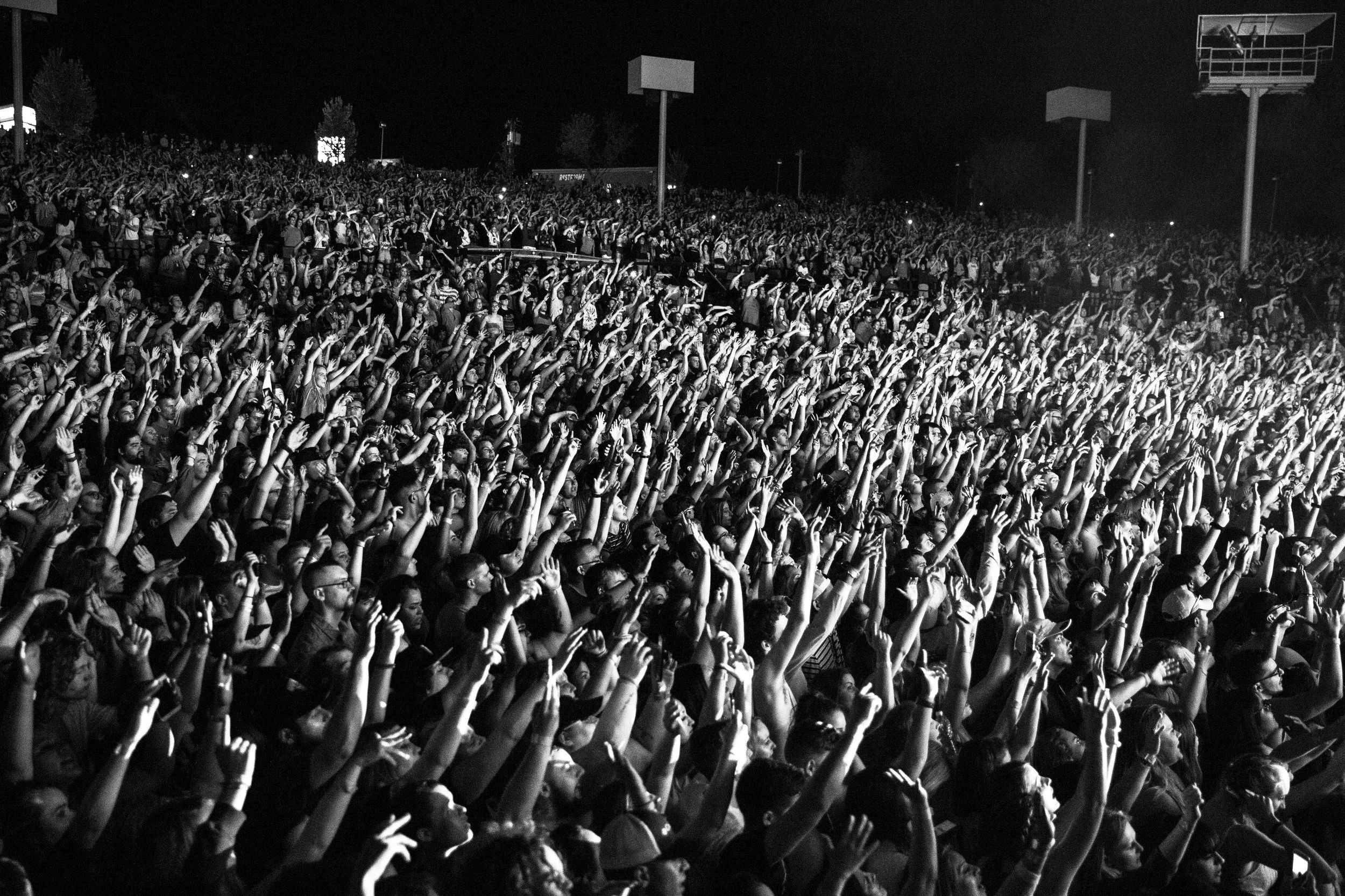 Thousands of music festival attendees gathered together with their hands raised during a live performance.