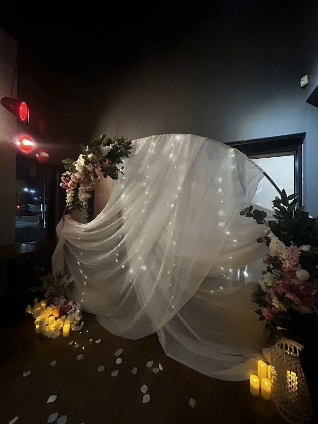 Wedding arch decorated with white flowing fabric, fairy lights, and floral arrangements, with candles and flower petals on the floor.