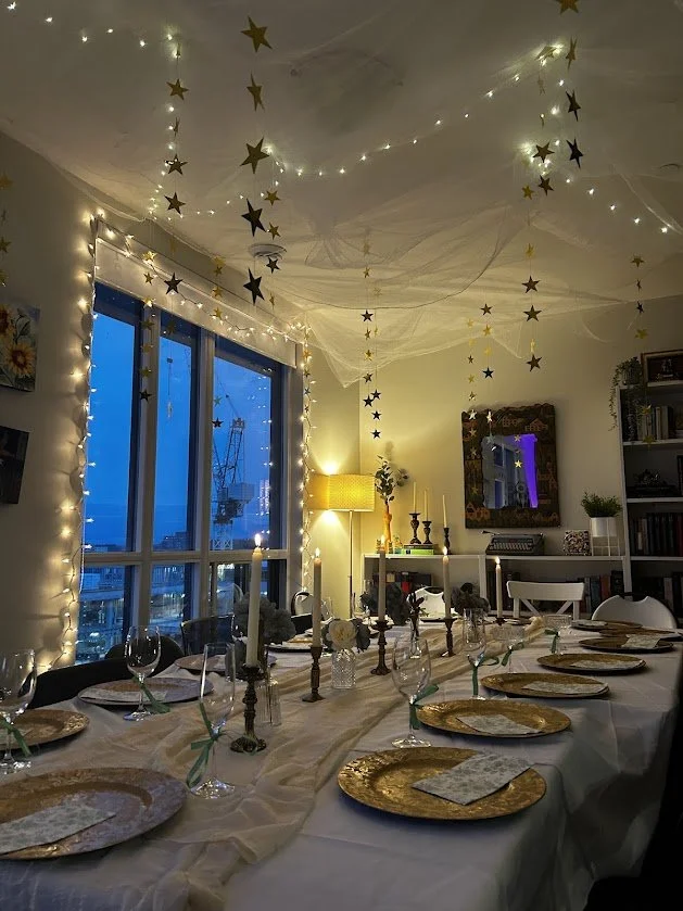 A dining room decorated with string lights and hanging star ornaments on the ceiling. The table is set with gold chargers, wine glasses, napkins tied with ribbons, and candleholders with white candles. There is a window showing a cityscape at dusk, a
