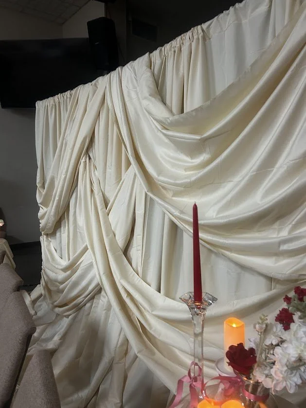 Cream-colored draped fabric backdrop with candles and flowers in the foreground, likely for a special event or wedding.