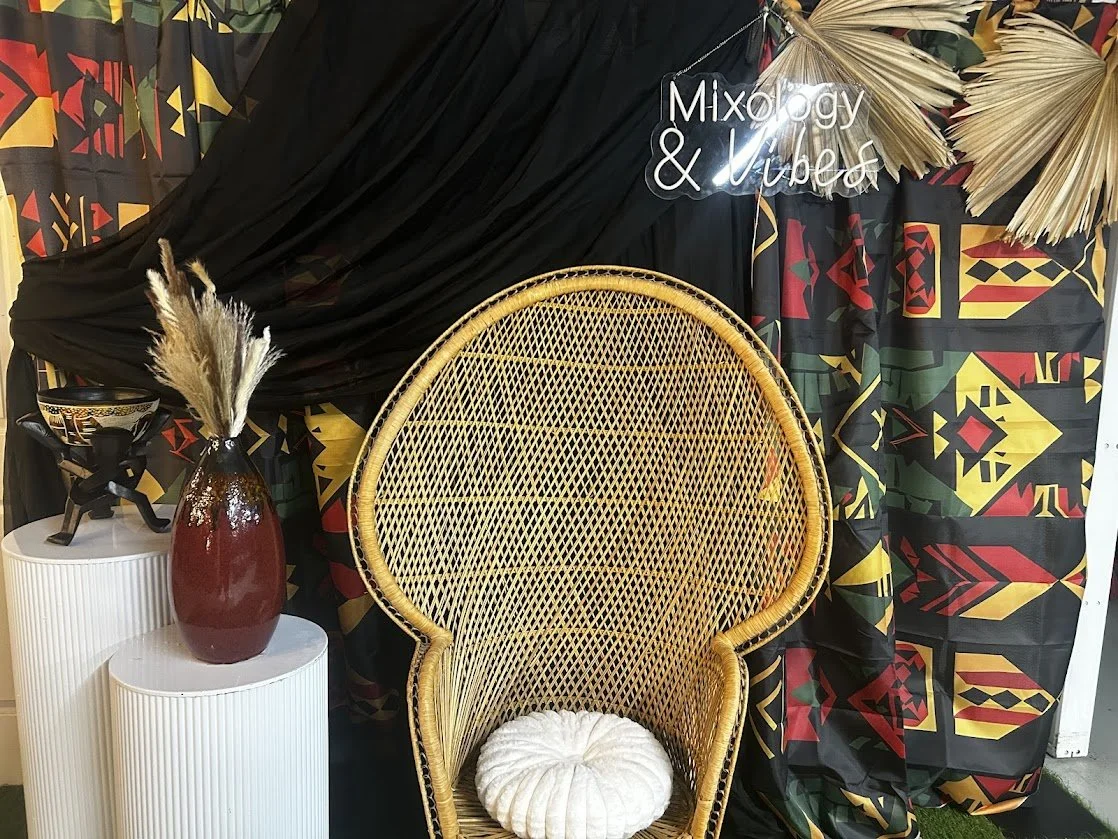 Decorative setup with a rattan chair with a white cushion, a red vase with pampas grass, patterned textiles, a black fabric backdrop, and a neon sign that says 'Mixology & Vibes'
