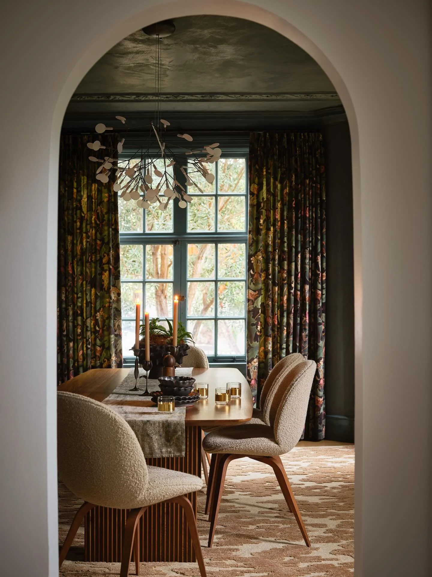 Dining in an elegant corner of the woods. 🌿 

Rich teal walls, a chandelier that mimics dappled raindrops, leafy printed curtains, walnut wood, cozy boucl&eacute; &mdash; every detail whispers nature, but make it a dinner party! This is the kind of 