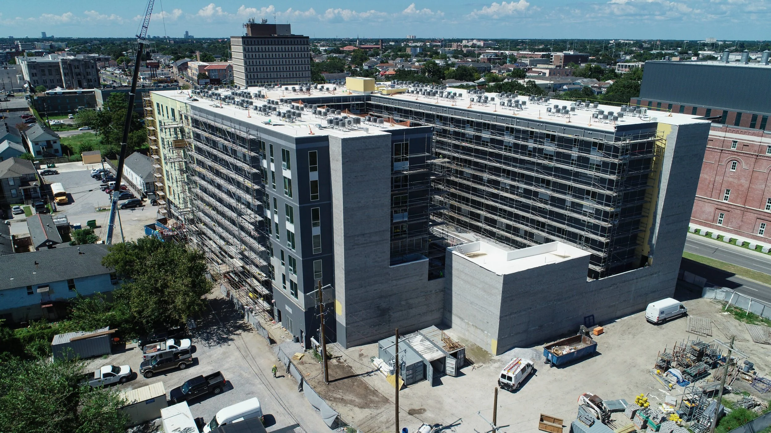 Aerial view of a multi-story building under construction with scaffolding, situated in an urban area with surrounding roads, smaller buildings, construction materials, and parked vehicles.