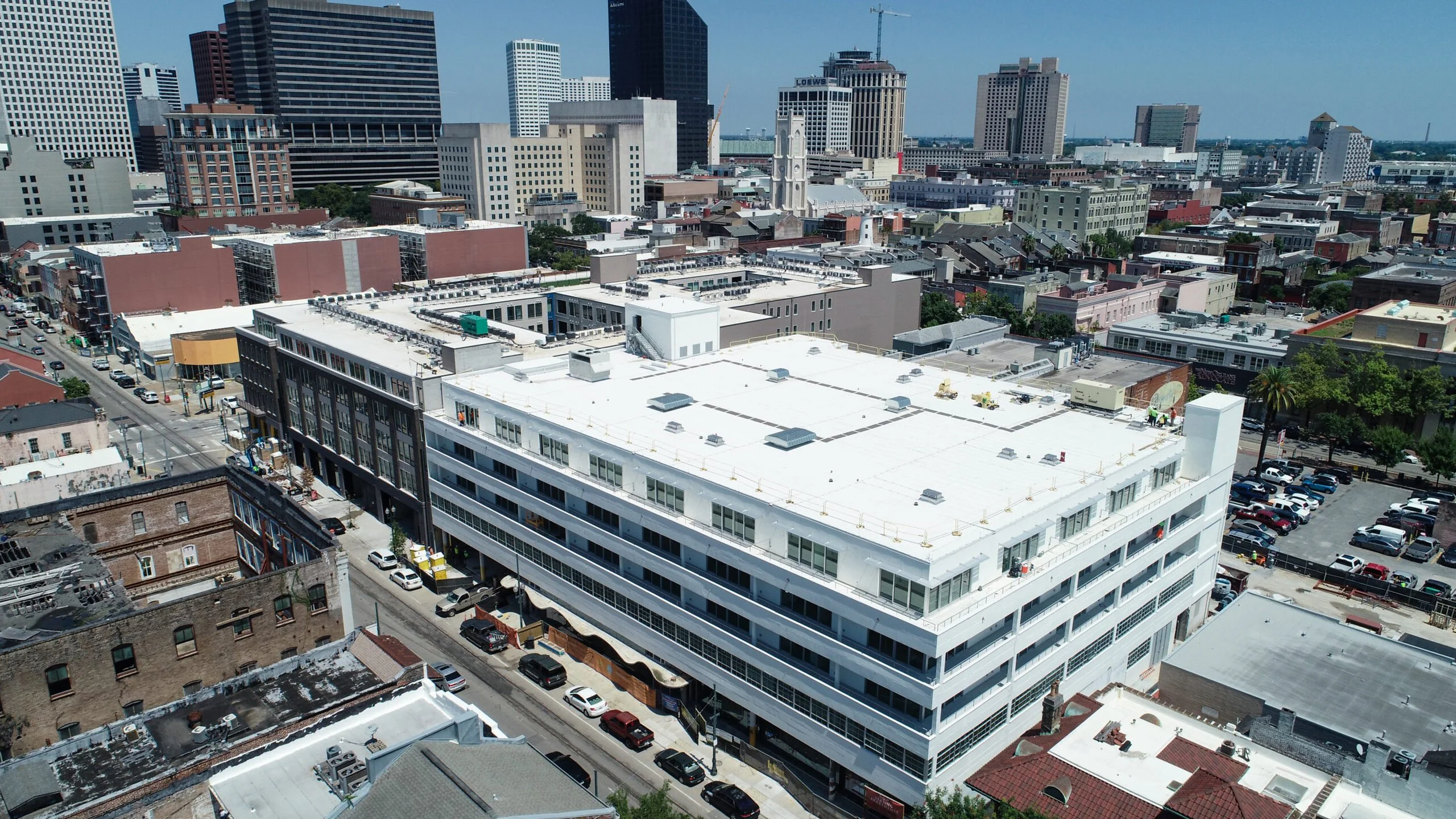 A cityscape featuring a large white building with many windows, surrounded by other buildings of varying heights and colors, with parking lots and streets in the foreground, under a clear blue sky.