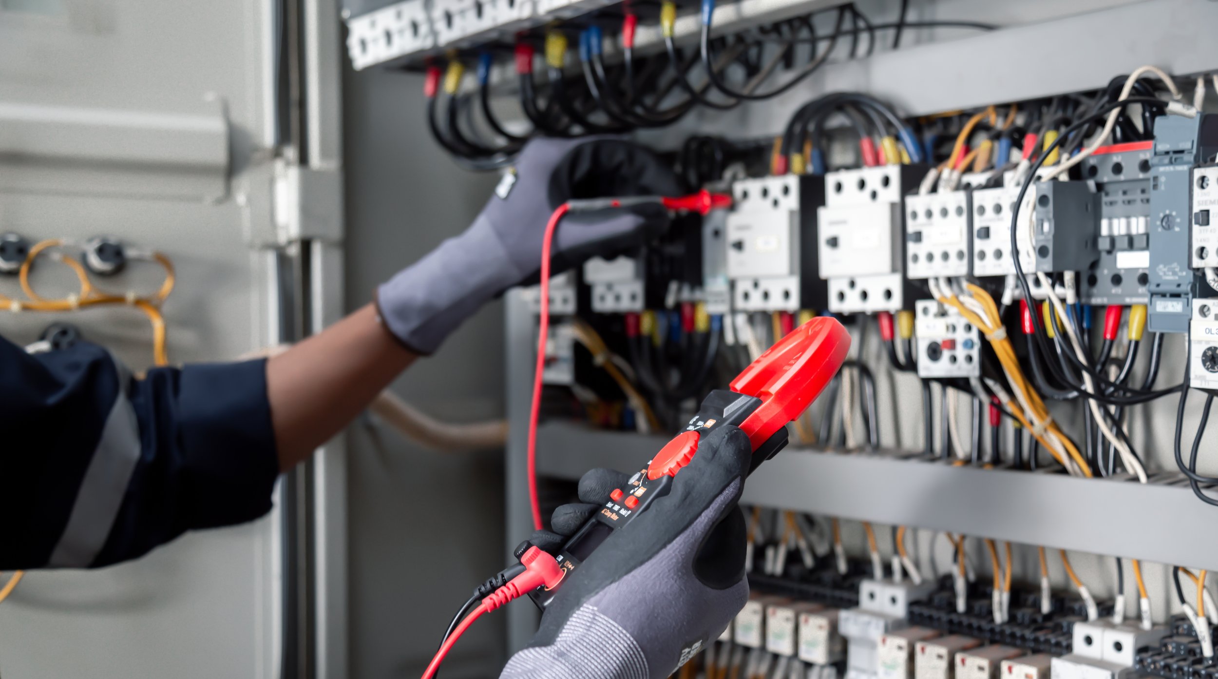 A technician wearing gloves uses a digital multimeter to test electrical wiring inside an electrical panel with various wires and components.