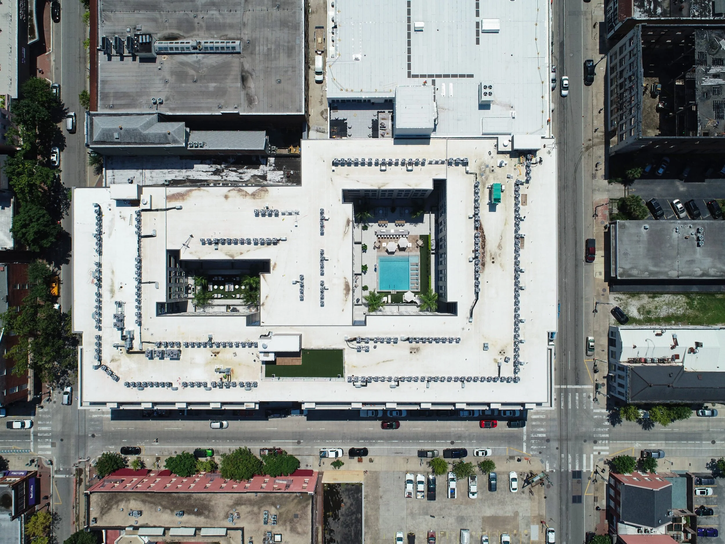 An aerial view of a city building with a central swimming pool on the rooftop surrounded by lounge chairs and umbrellas, with streets and parked cars around it.