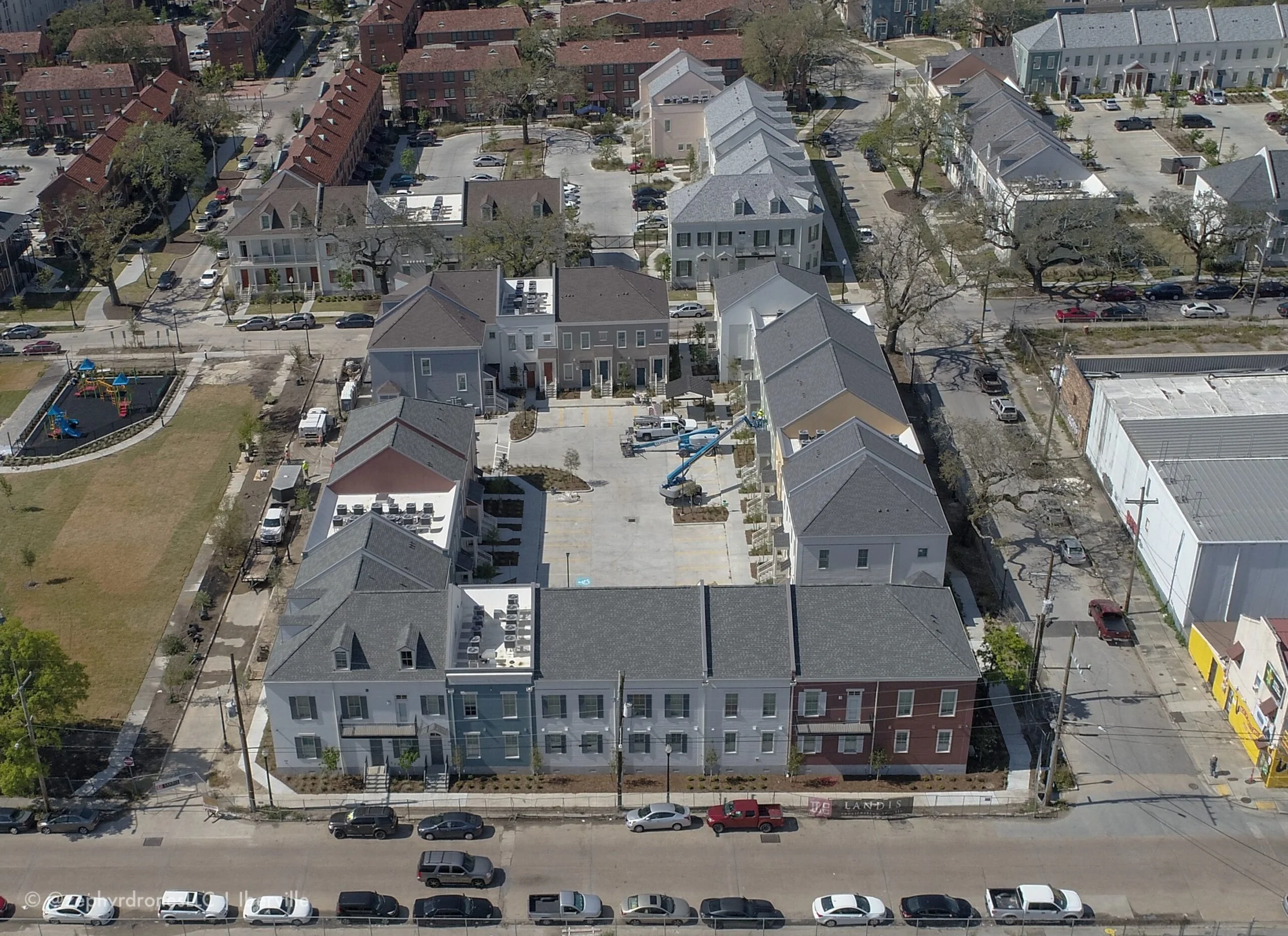 An aerial view of a residential neighborhood showing a new apartment complex with multiple buildings, a central parking lot, a playground on the left, and surrounding houses and streets.