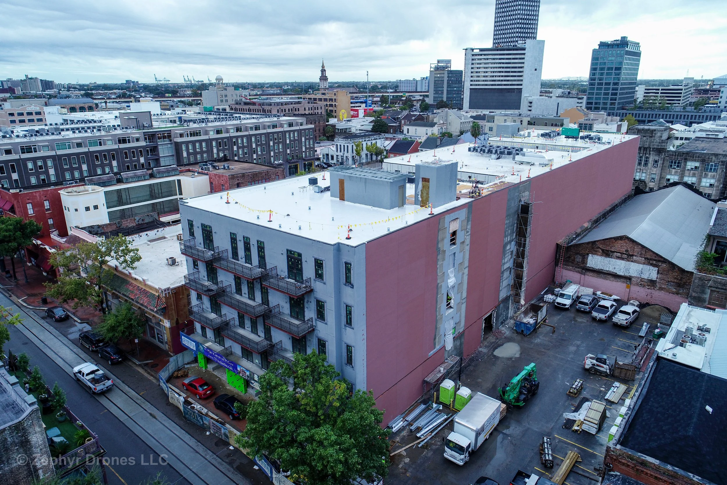 Aerial view of a construction site on a city block with multi-story buildings, some under renovation, and a parking lot with trucks and equipment, with a skyline of high-rise buildings in the background under cloudy skies.