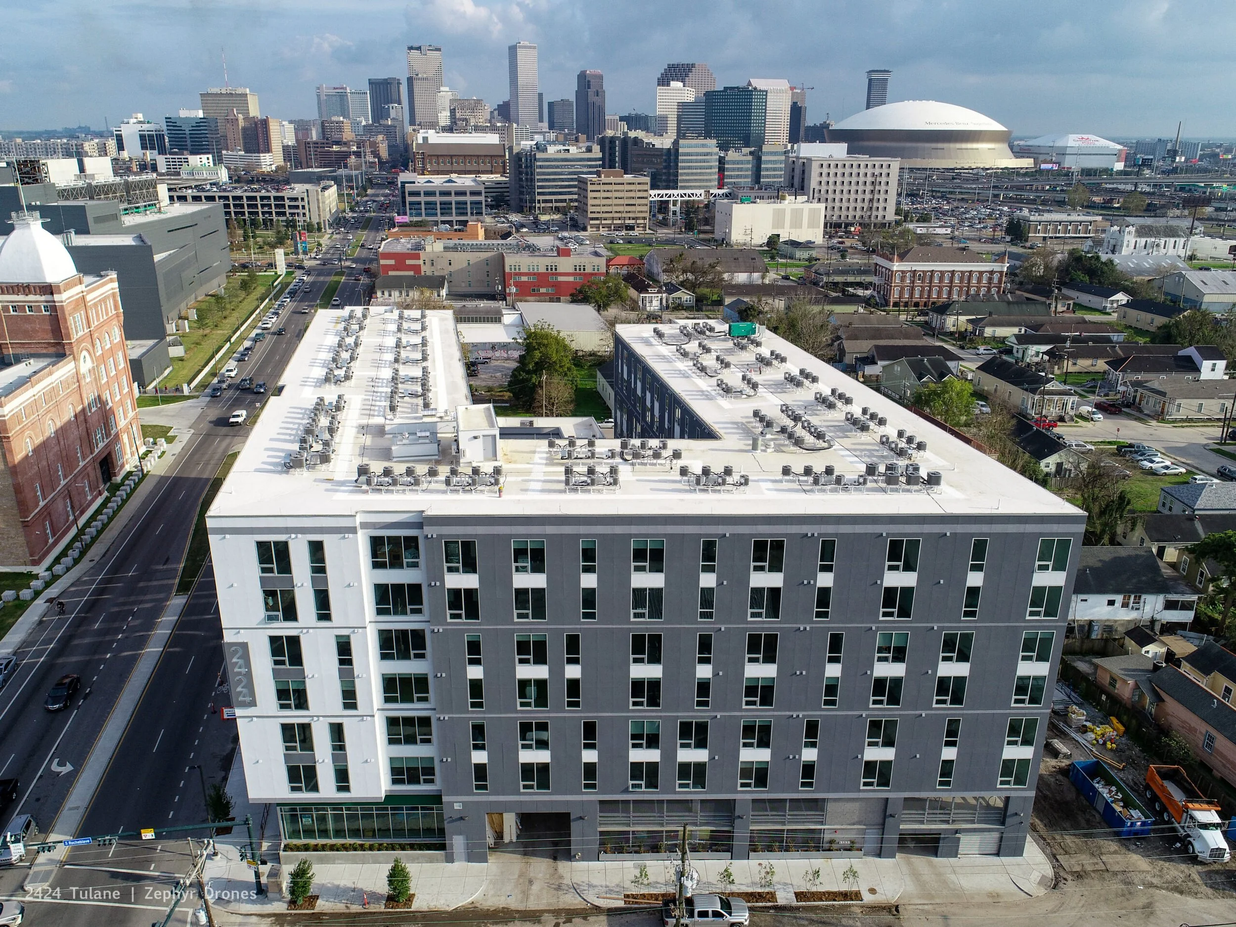 Aerial view of a cityscape showing a modern multi-story building with a flat roof, with numerous HVAC units on top, surrounded by streets, older buildings, and tall skyscrapers in the background.
