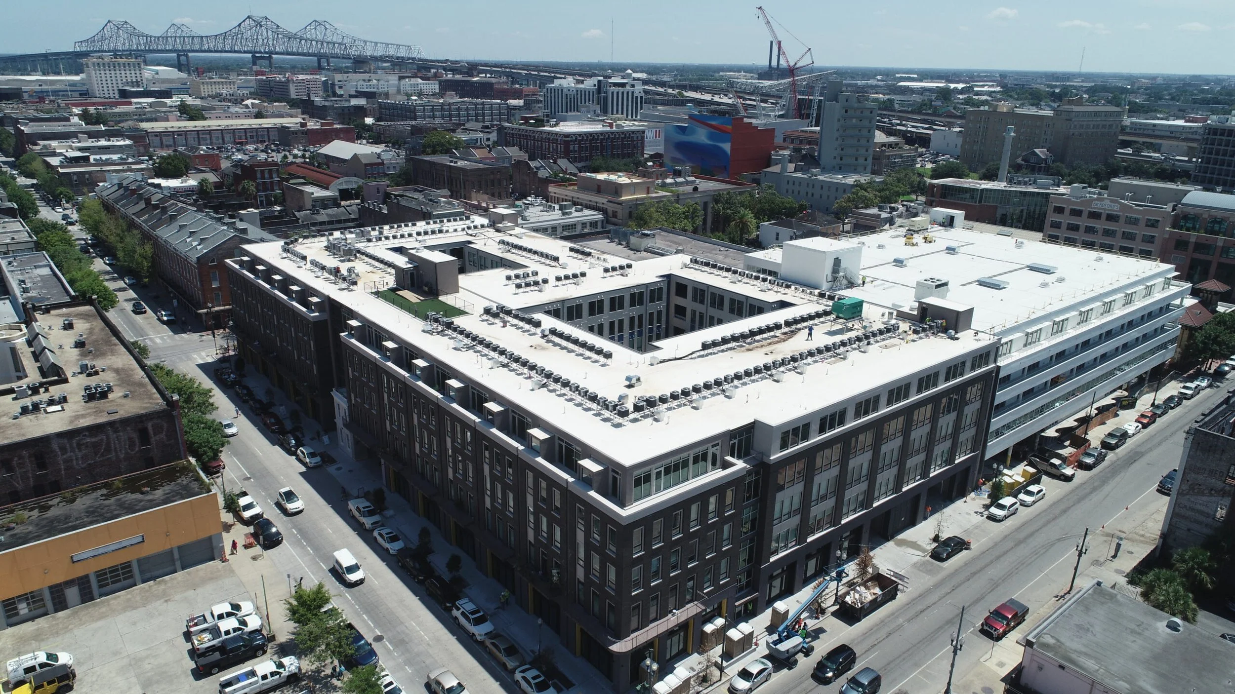 Aerial view of a cityscape with a large modern building in the foreground, surrounded by streets filled with parked cars, and a bridge and cranes visible in the background under a partly cloudy sky.