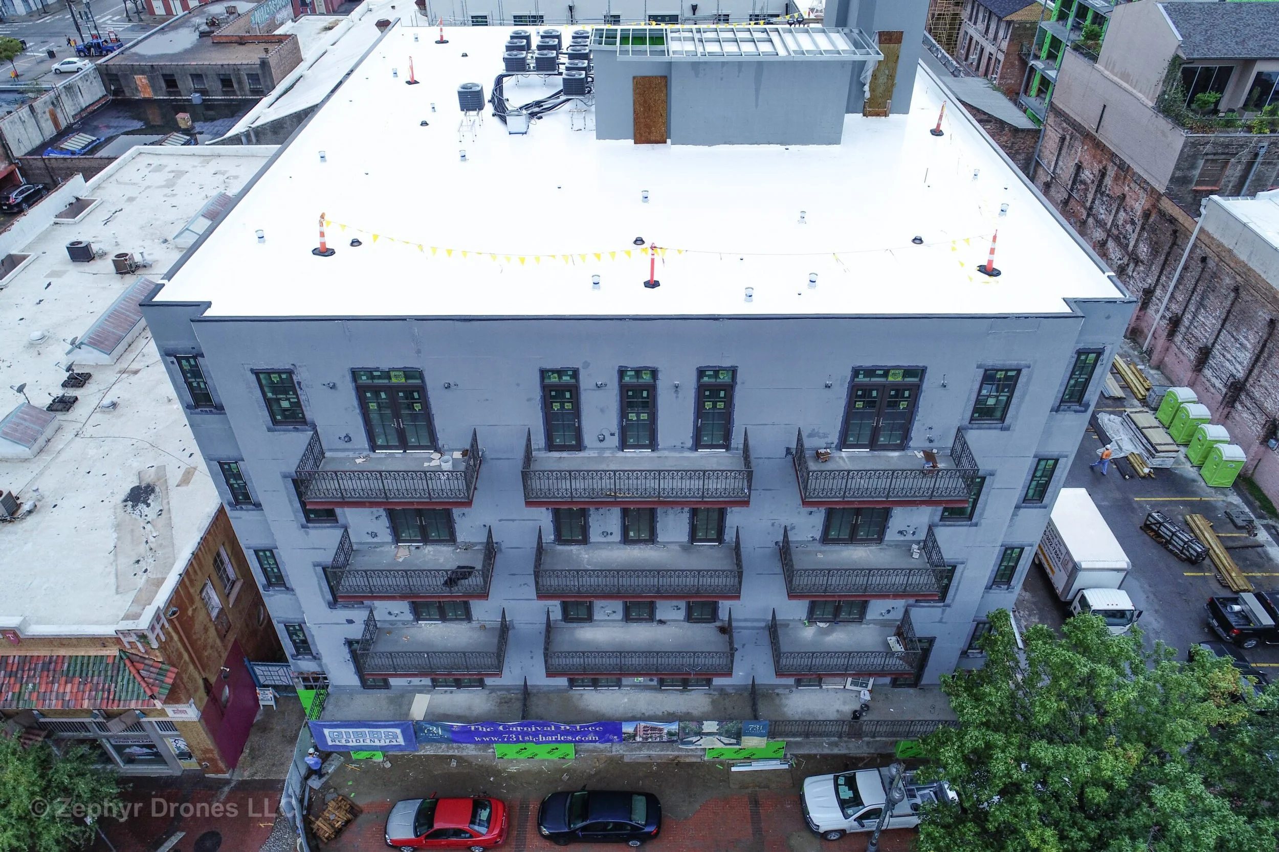 An aerial view of a four-story building with balconies, situated in an urban area. The building has a flat roof with construction cones and equipment, and neighboring buildings and a parking lot are visible.